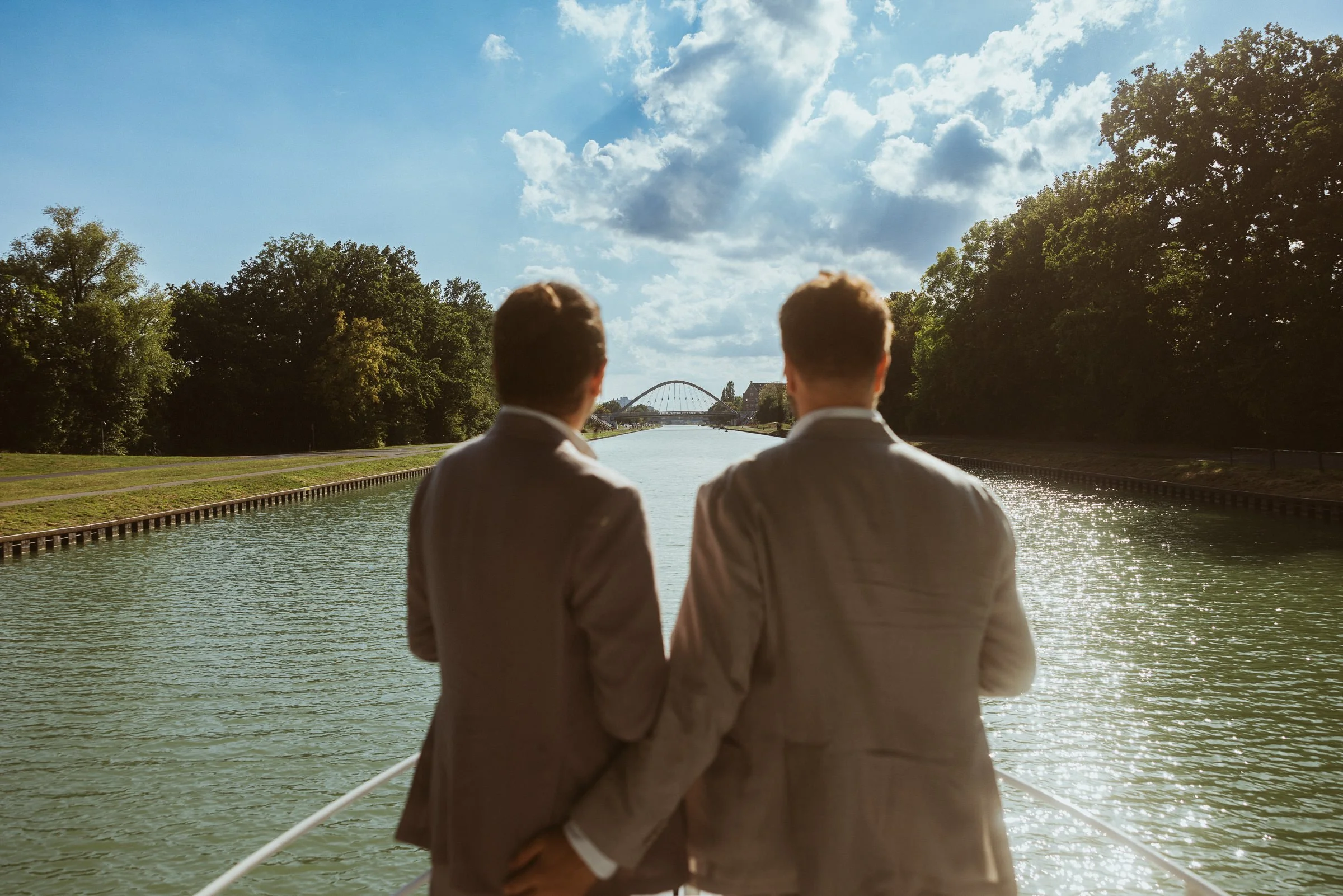 Zwei Männer in Anzügen stehen am Fluss und schauen auf eine Brücke im Hintergrund, umgeben von Bäumen und einer bewölkten blauen Himmel
