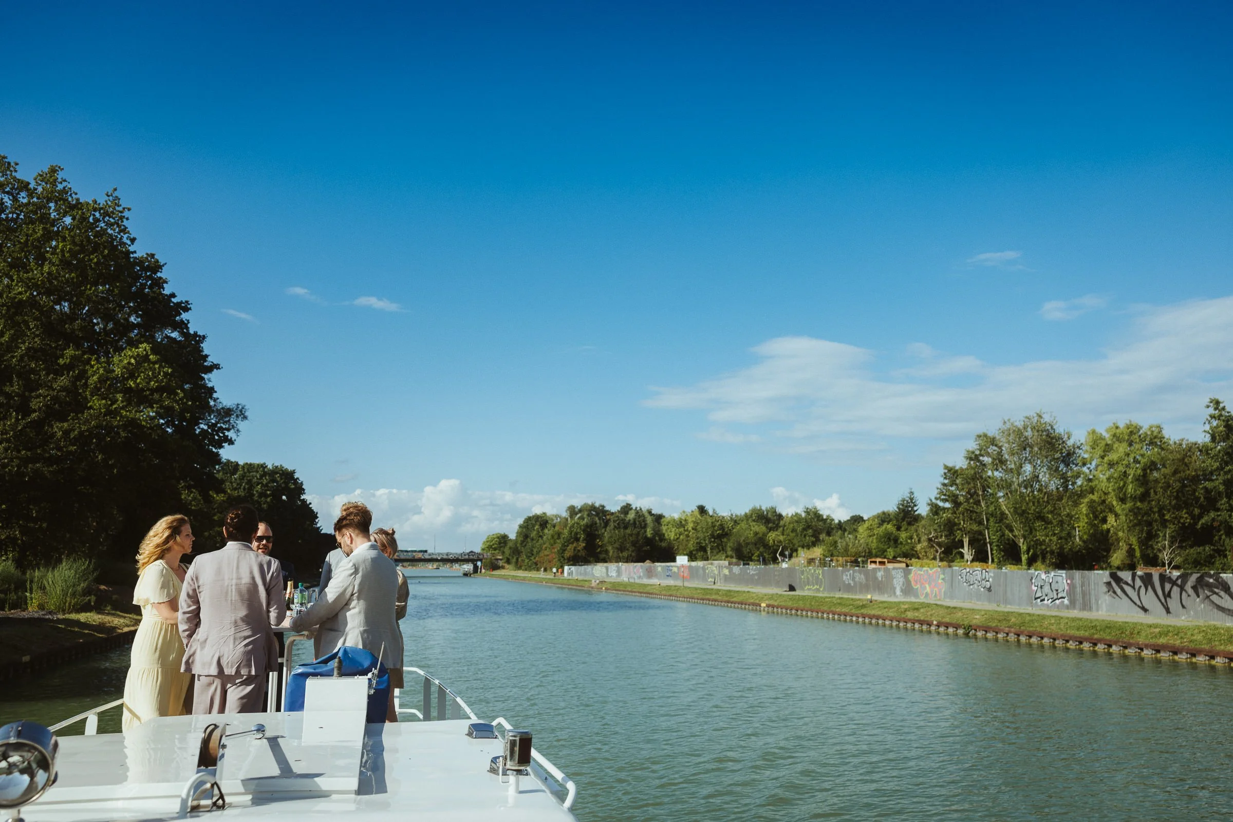 Menschen auf einem Boot entlang eines Flusses mit einer grünen Uferpromenade und Graffiti an der Mauer, im Hintergrund Bäume und ein blauer Himmel.