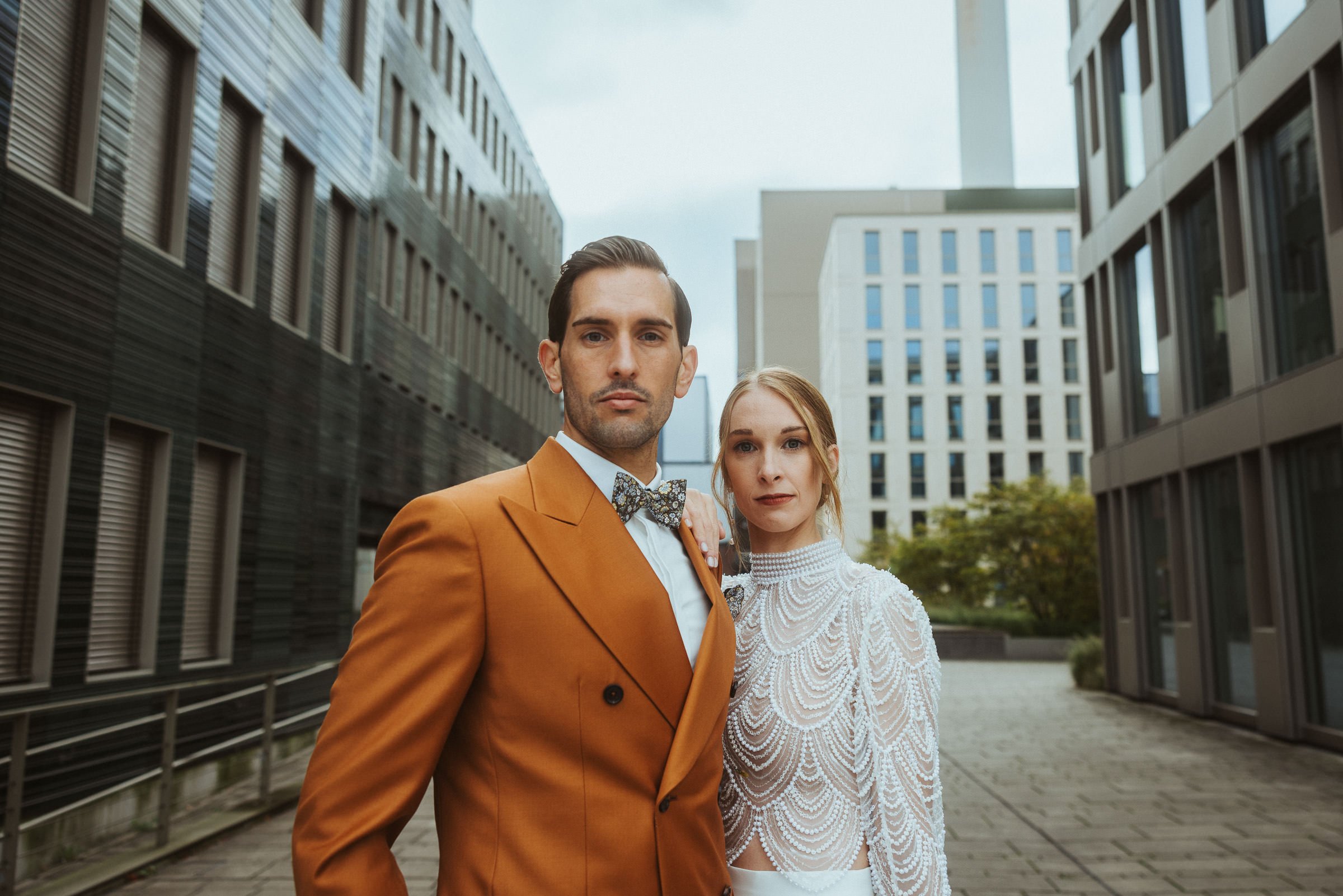 Ein Paar in formeller Kleidung steht vor modernen Bürogebäuden, der Mann trägt einen braunen Anzug mit Fliege, die Frau trägt ein weißes, detailliertes Kleid.