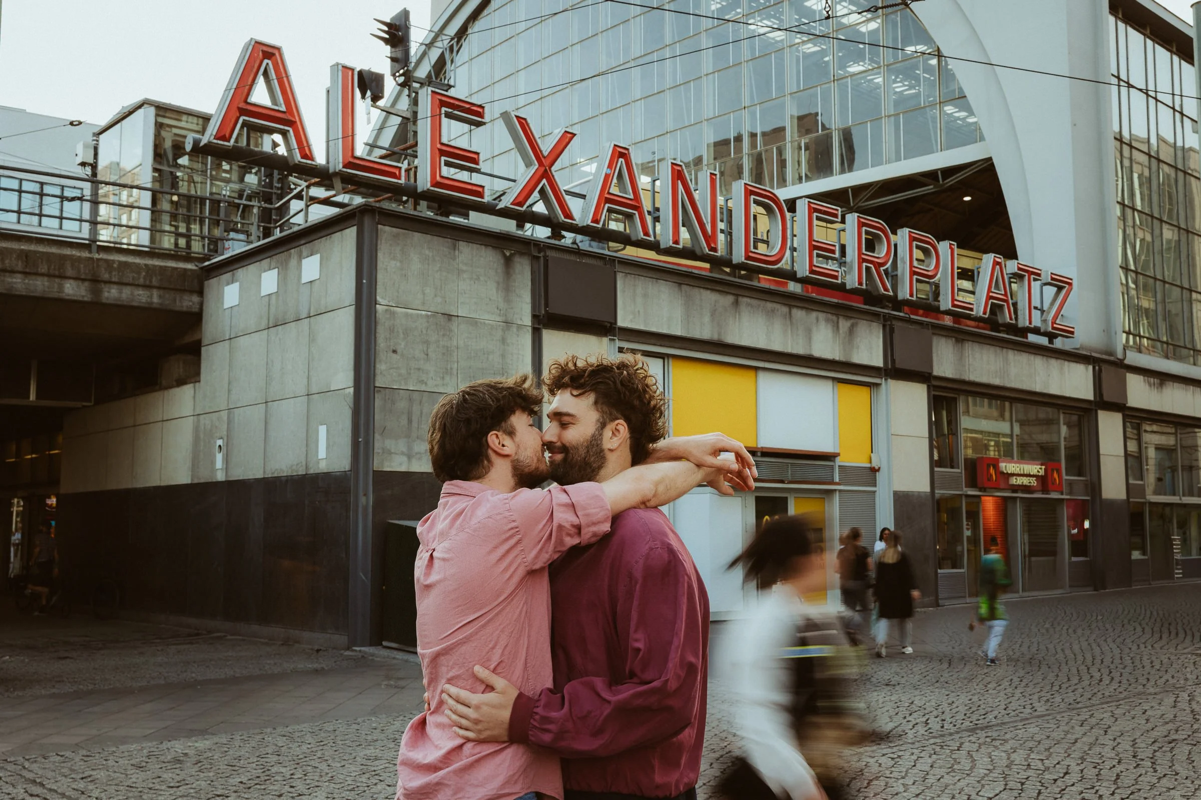 Zwei Männer umarmen sich auf einer Stadtstraße, im Hintergrund das Gebäude Alexanderplatz mit rotem beleuchteten Schriftzug. Es gibt Bewegungsunschärfe bei den vorbeigehenden Personen.