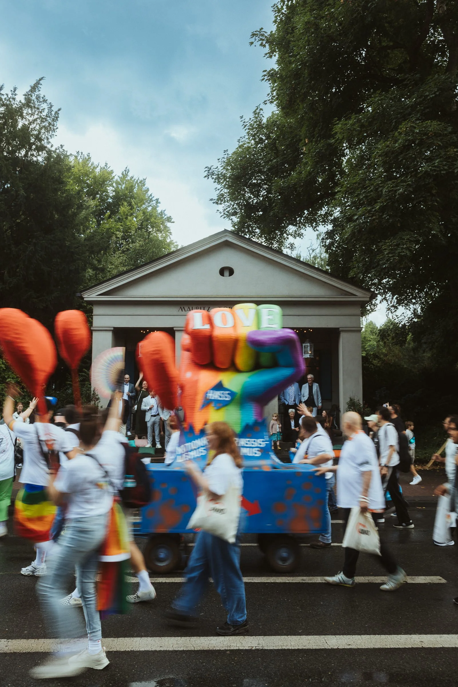 Menschen bei einer Parade oder Demonstration, im Vordergrund eine große, bunte Ballonhand mit den Buchstaben L, O, V, E, auf einem Wagen, im Hintergrund ein Gebäude mit Säulen und umgeben von Bäumen, über dem Gebäude steht MAURITZ.