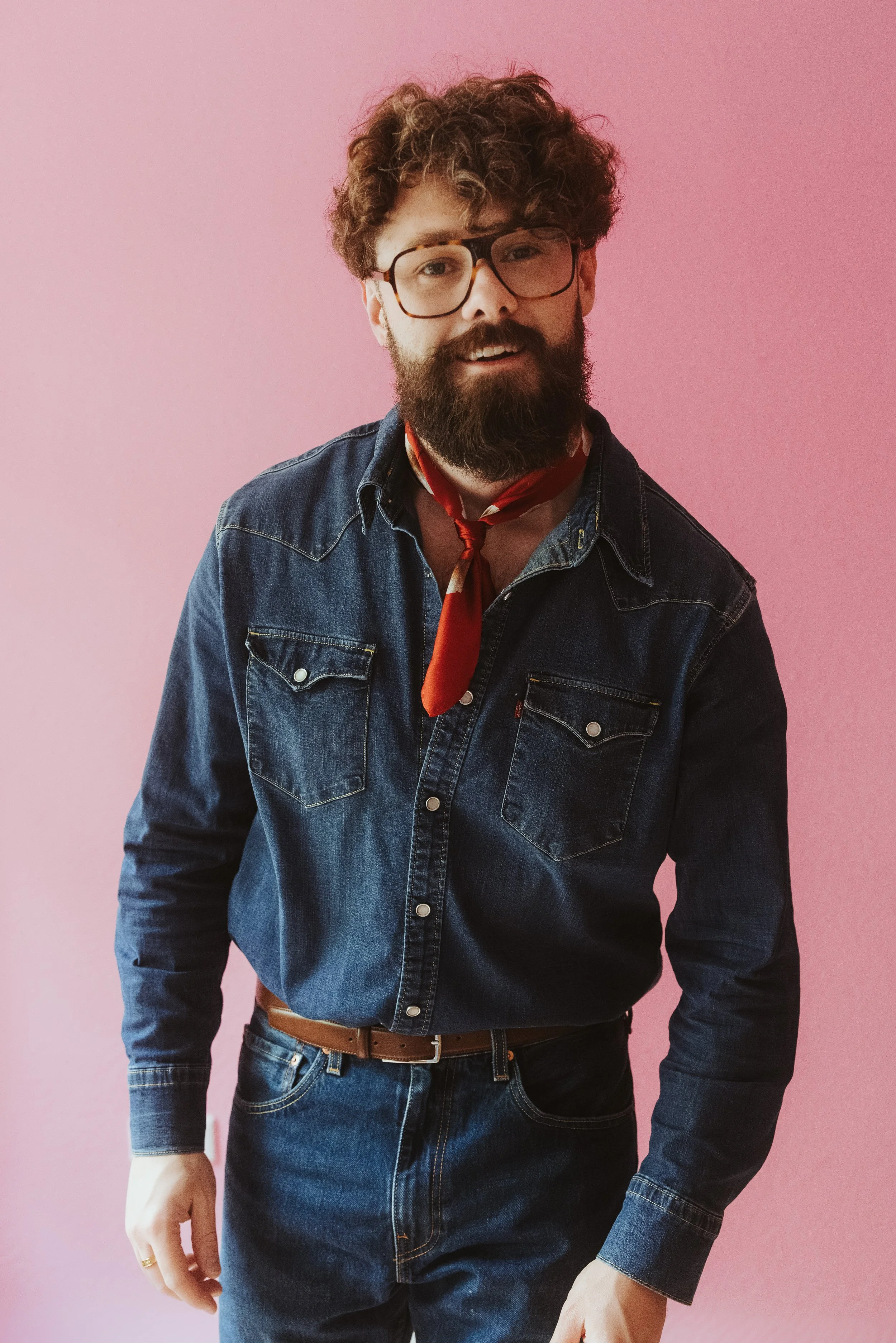 Ein junger Mann mit Locken, Brille und Bart trägt eine schwarze Jeansjacke, einen roten Schal und eine braune Gürtelhose, steht vor einem pinkfarbenen Hintergrund.