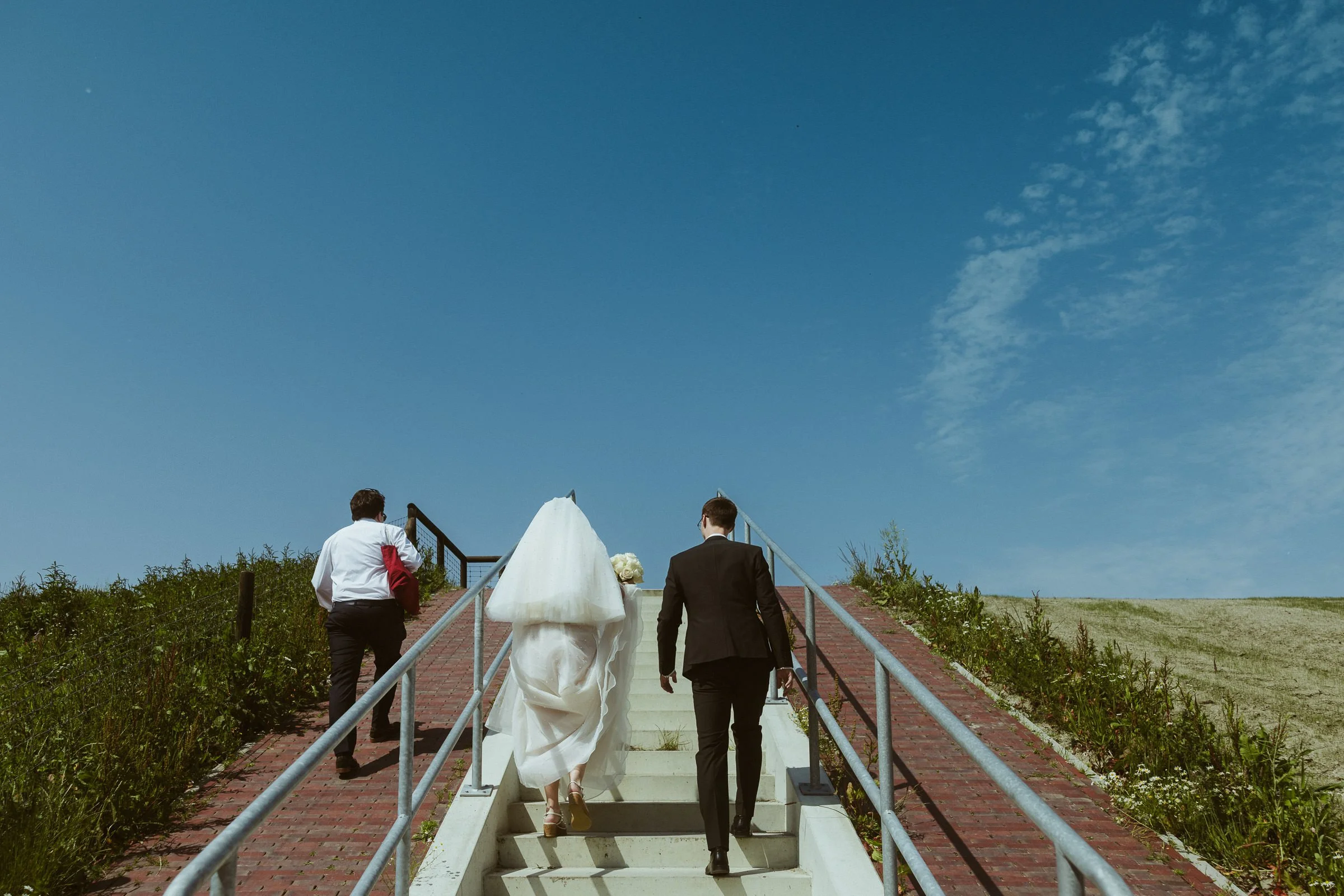 Zwei Menschen in formeller Kleidung laufen eine weiße Treppe hinauf, eine davon trägt ein Brautkleid, die andere einen schwarzen Anzug, unter einem blauen Himmel mit wenigen Wolken.