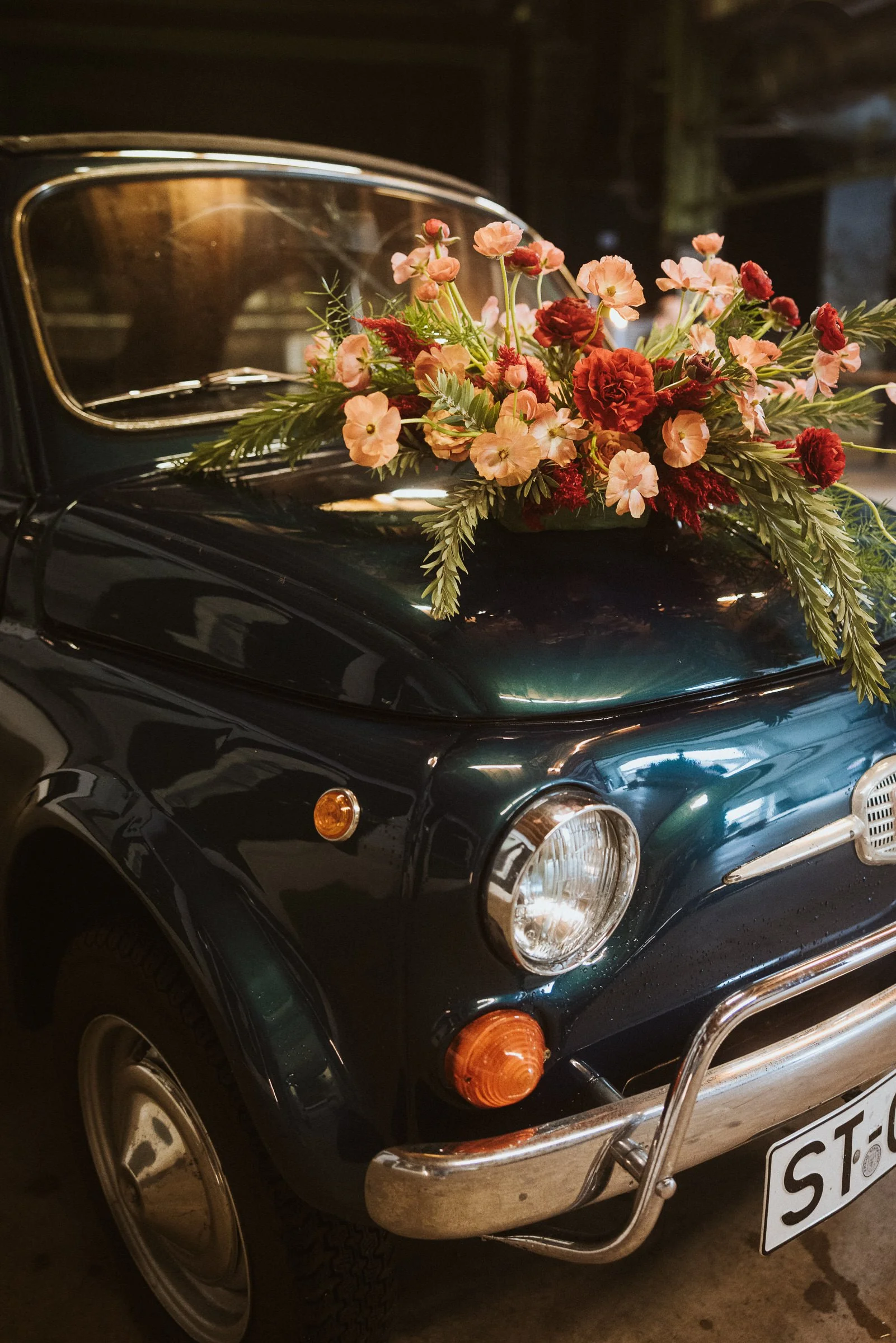 Ein klassischer schwarzer Oldtimer mit einem Blumenstrauß aus roten und pinken Blumen auf der Motorhaube. Das Auto ist in einem Parkhaus oder ähnlichem Ort geparkt.