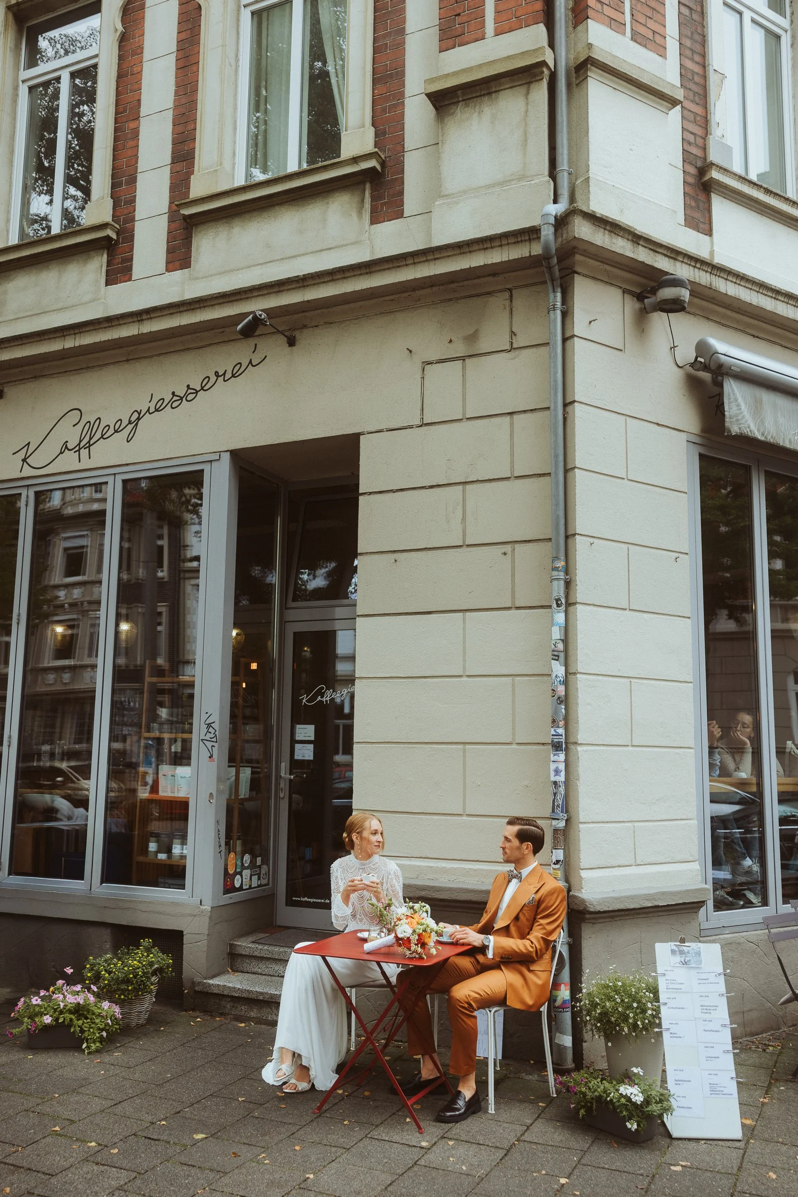 Ein Pärchen sitzt an einem kleinen Tisch vor einem Café, das Café-Kaffeehaus, mit einem Blumenstrauß auf dem Tisch. Das Paar trägt formelle Kleidung, er in einem braunen Anzug und sie in einem weißen Kleid. Im Hintergrund sieht man das Café-Geschäft 