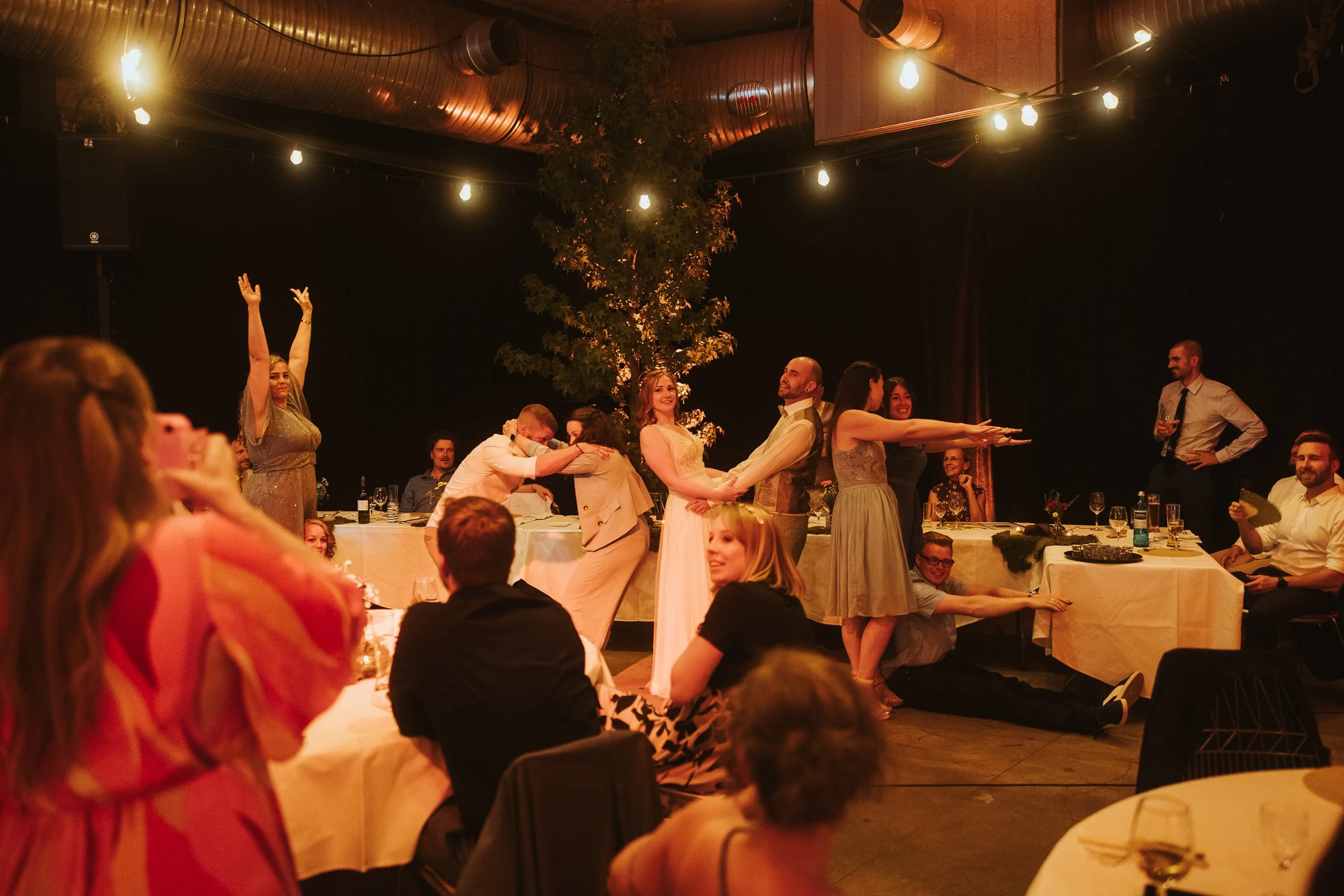 Menschen feiern eine Hochzeit im festlich gedeckten Raum mit Lichterketten, einem Baum in der Mitte und mehreren Paaren, die tanzen und lachen.
