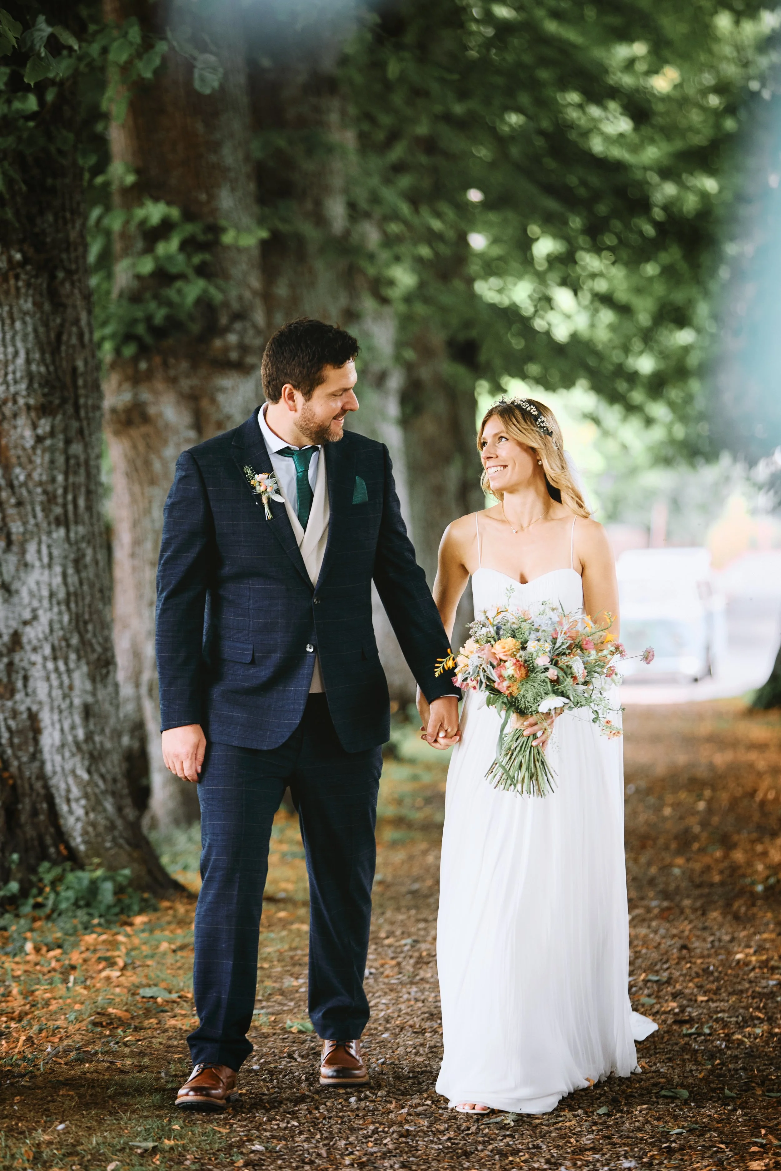 A bride and groom walking hand in hand outdoors, surrounded by large trees with green leaves, smiling at each other on their wedding day. The bride wears a white dress and holds a bouquet of flowers, while the groom wears a dark suit and tie.
