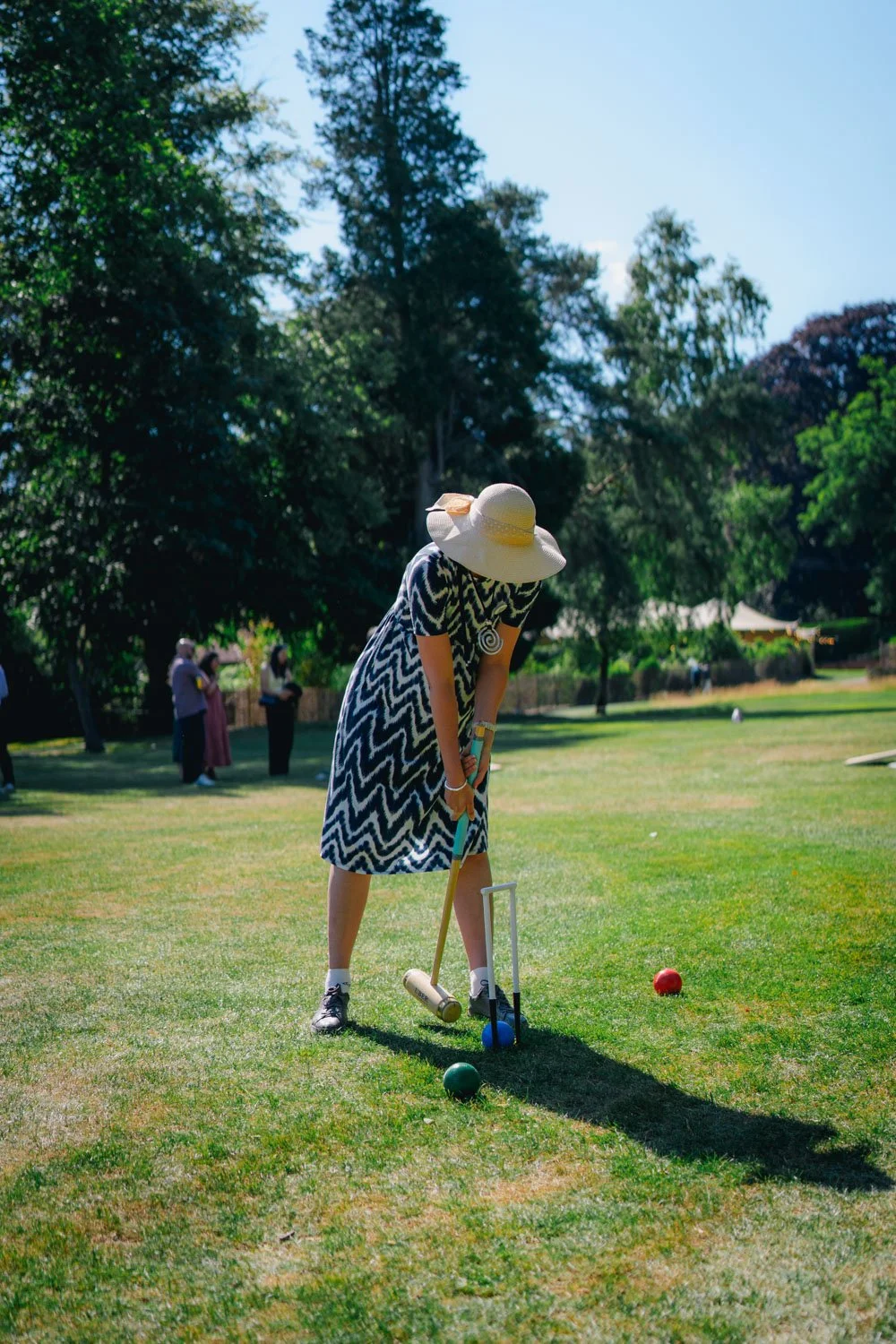 A woman playing croquet outdoors on a sunny day, wearing a wide-brimmed hat and a black-and-white zigzag dress, bent over aiming her mallet at a blue ball, with other players and trees in the background.