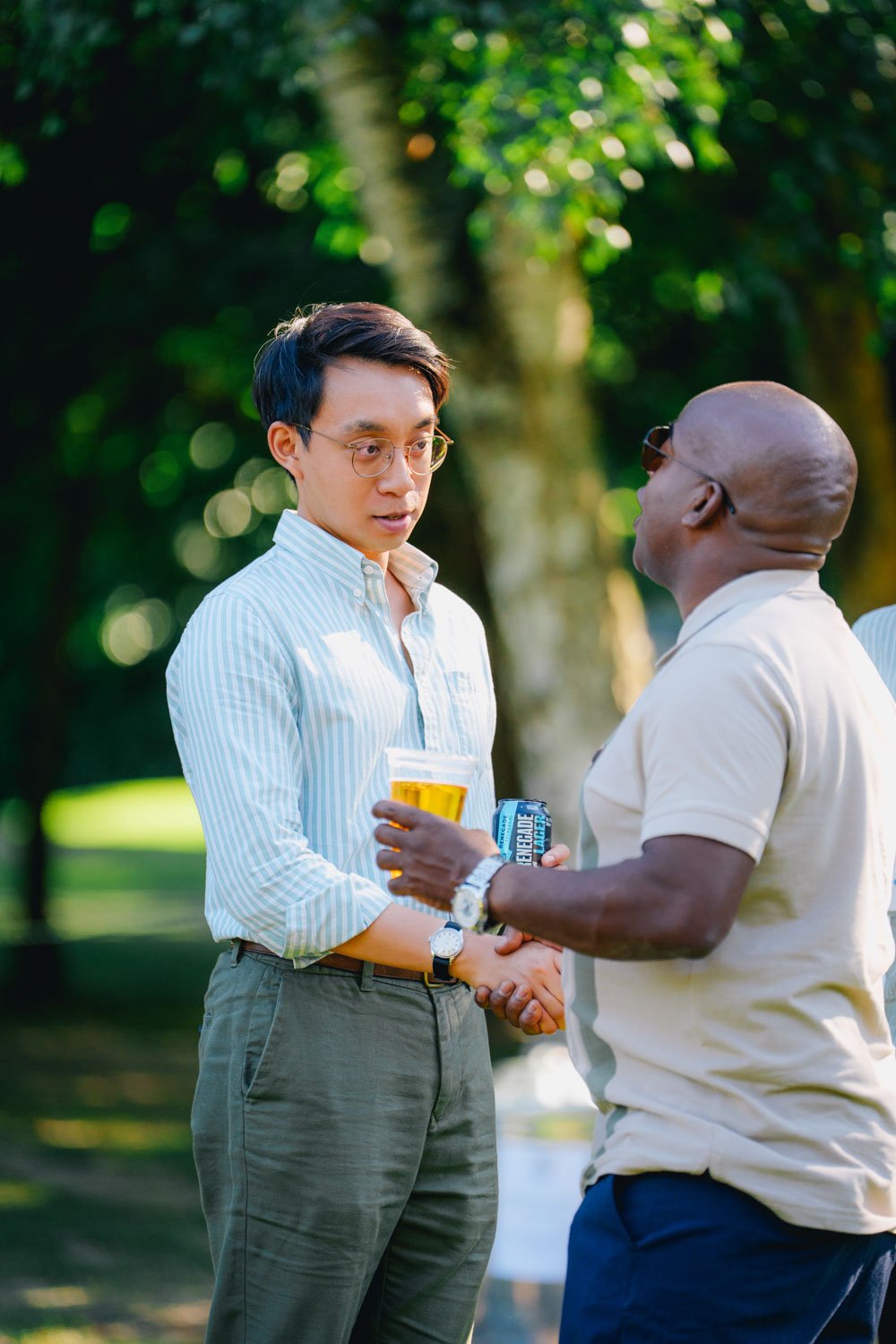 Two men shaking hands outdoors, one holding a drink and a beer, with trees and sunlight in the background.