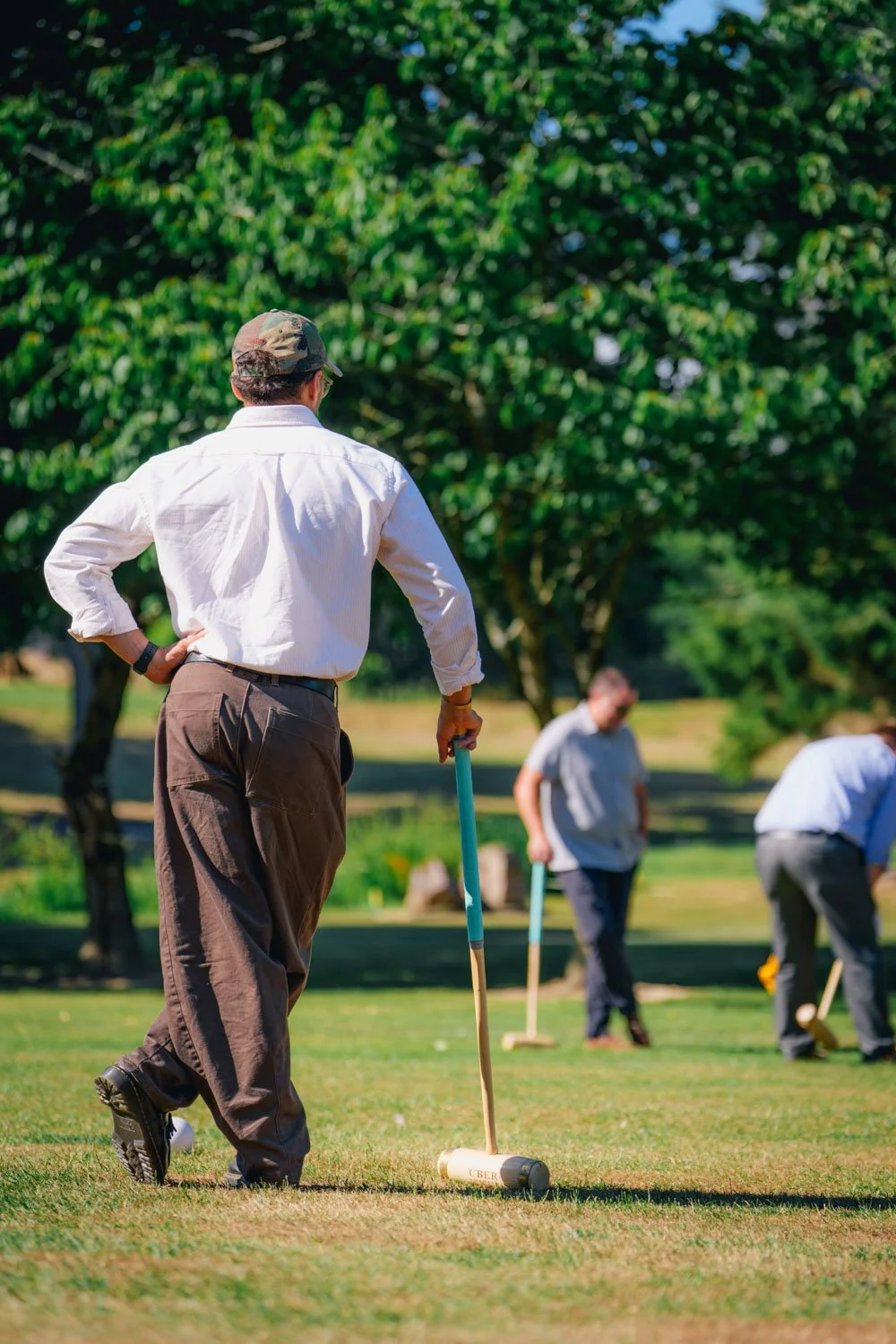 Man with white shirt and cap preparing to strike a croquet ball on a grassy lawn with others in the background.