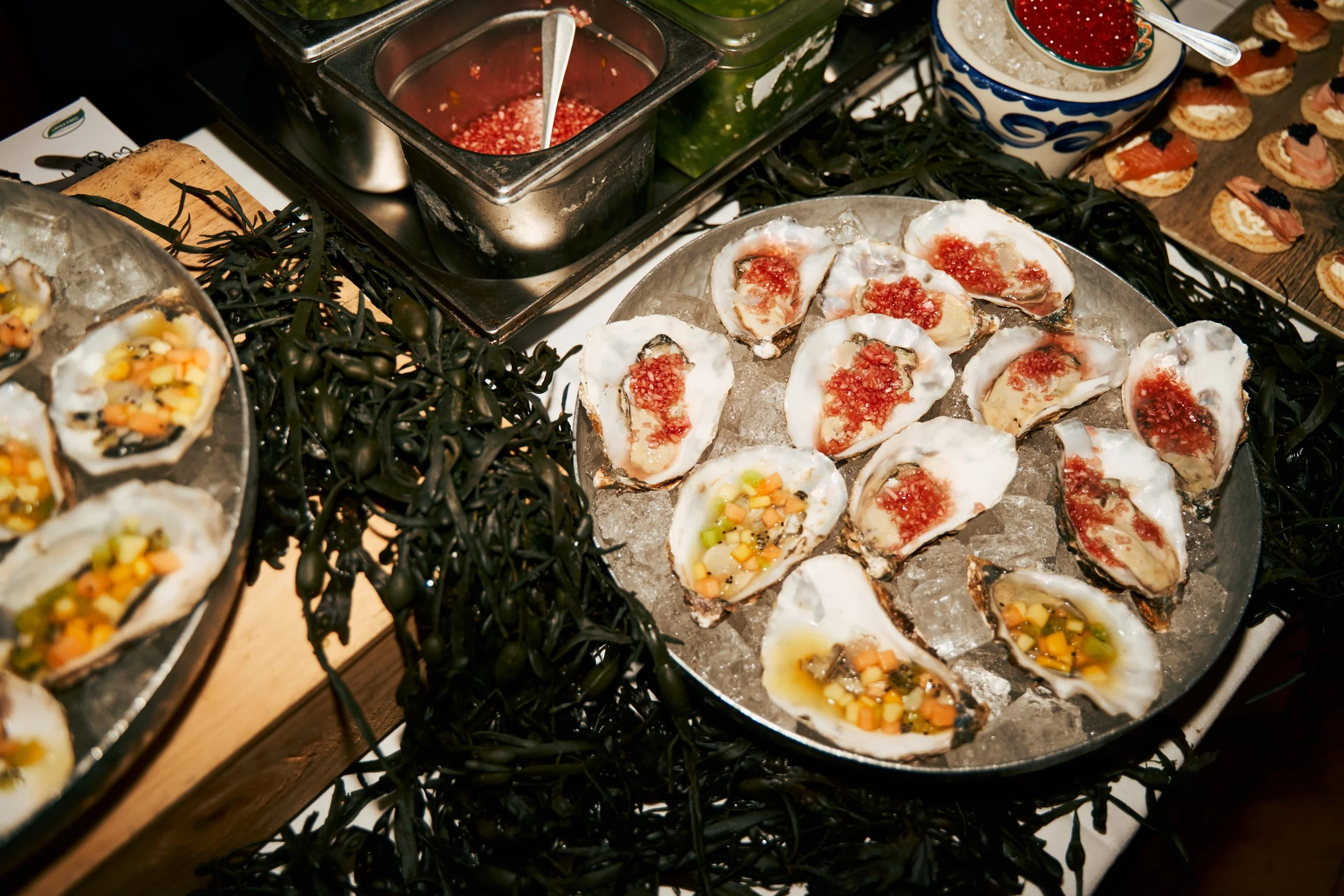Oysters on a bed of ice with chopped vegetables and red sauce, surrounded by greenery and other appetizers at a buffet.