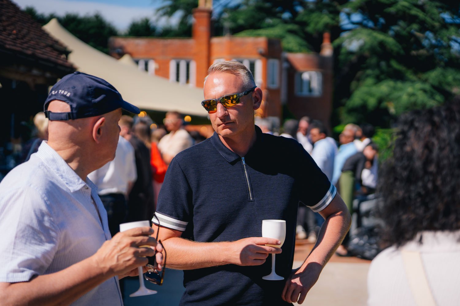 People socializing outdoors at a gathering, holding white wine glasses, under sunlight with a backdrop of trees and buildings.
