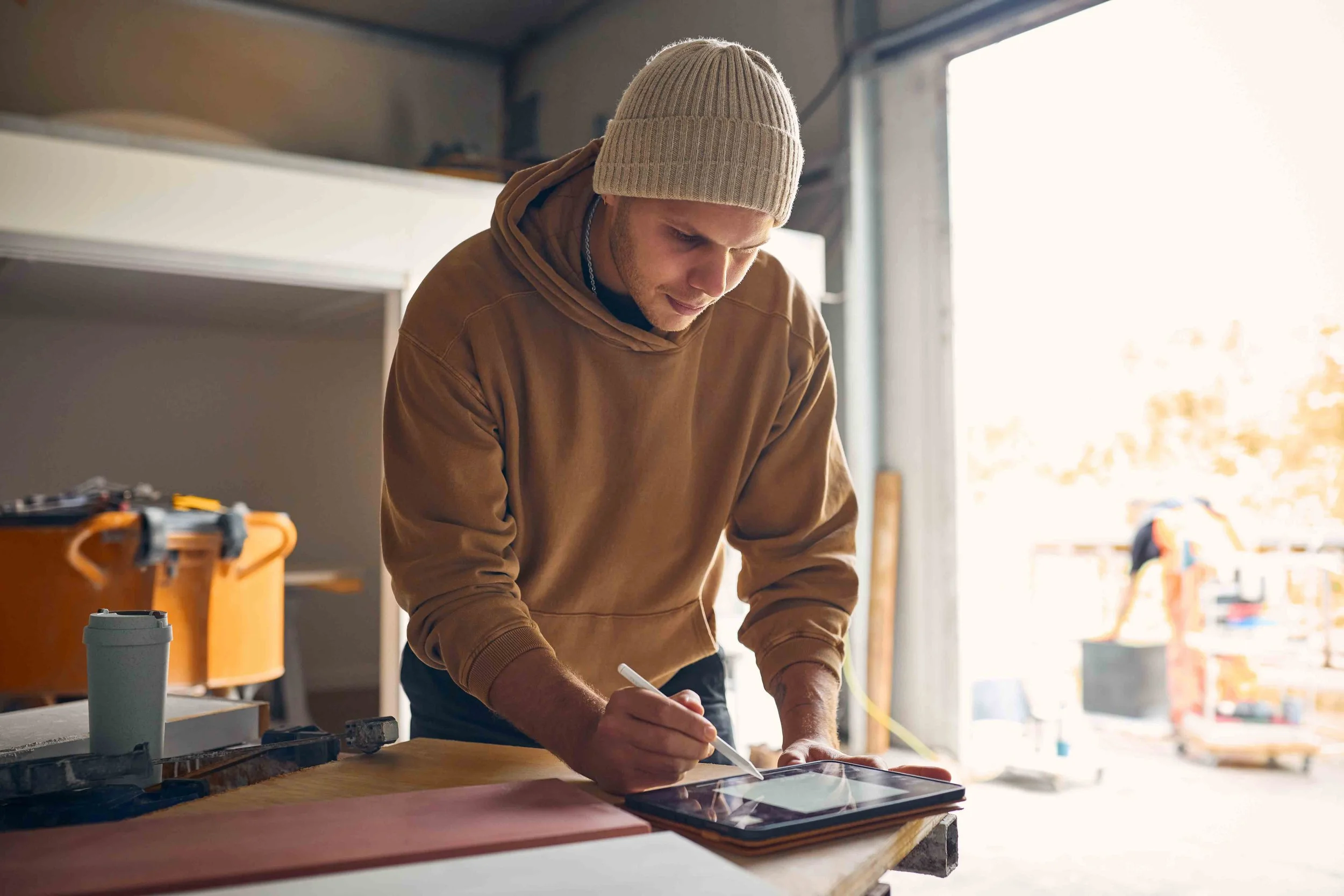 A man wearing a beige knit beanie and brown hoodie working on a tablet or device in a workshop with natural light coming through large windows.