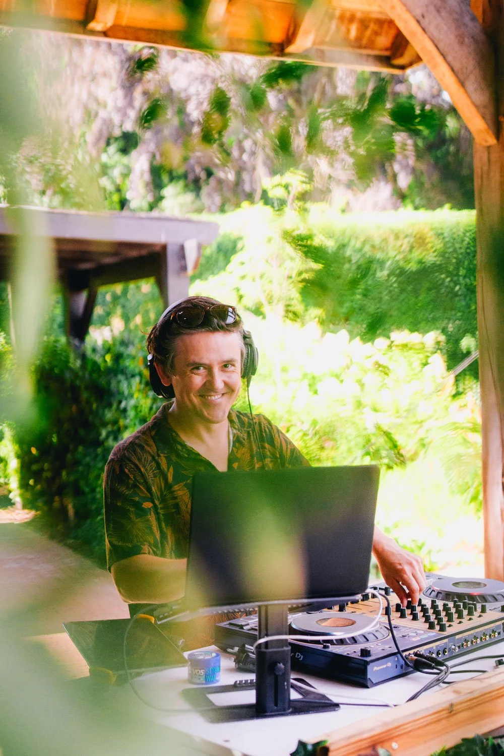 A smiling male DJ in tropical outdoor setting behind DJ equipment, wearing headphones, with lush greenery in background.