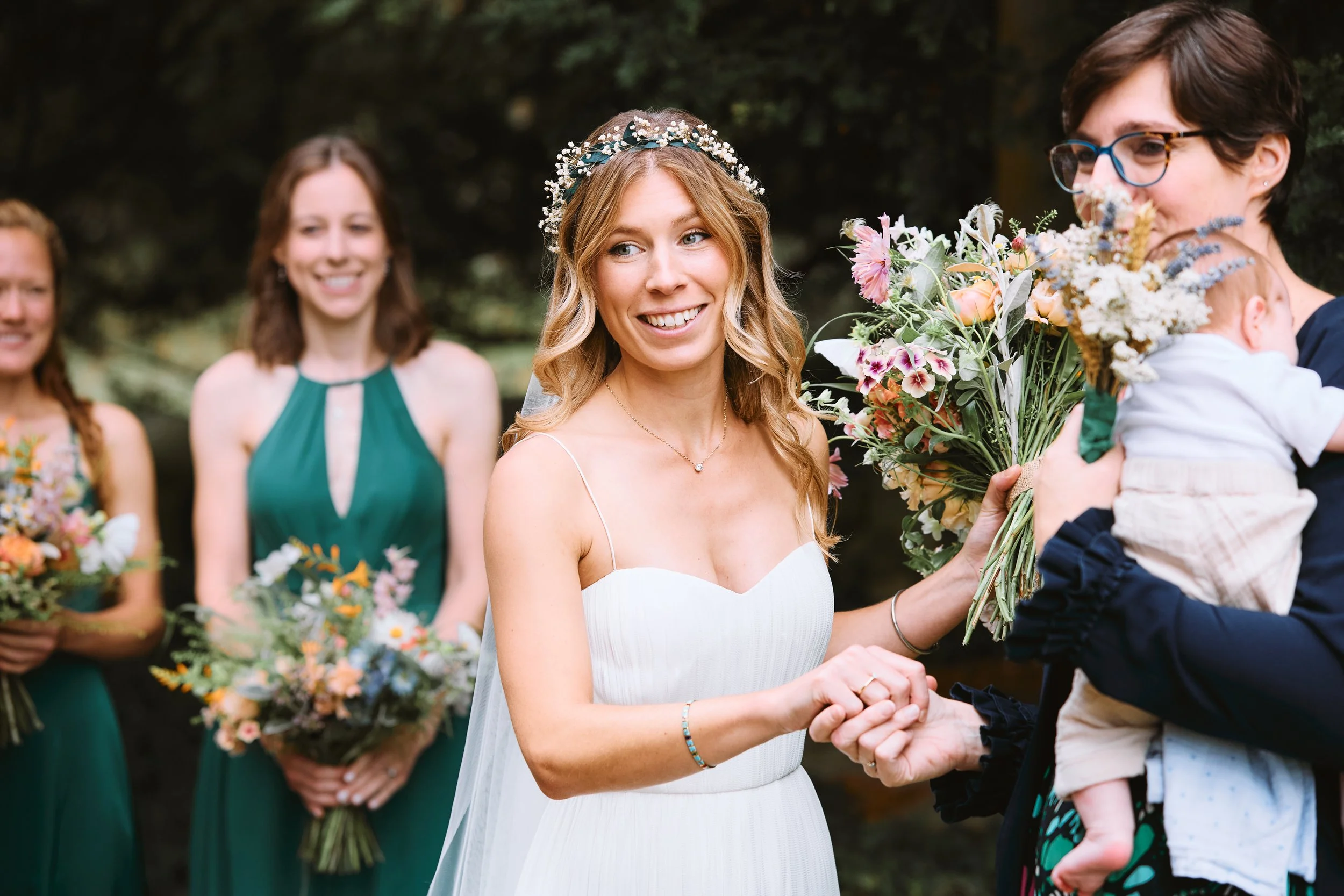 A wedding ceremony outdoors with a bride in a white dress shaking hands with a woman holding a baby, surrounded by bridesmaids holding bouquets of flowers, with trees in the background.