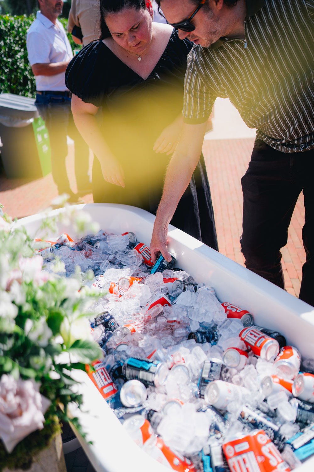 People are disposing of empty soda cans and bottles into a large white container filled with ice at an outdoor event.