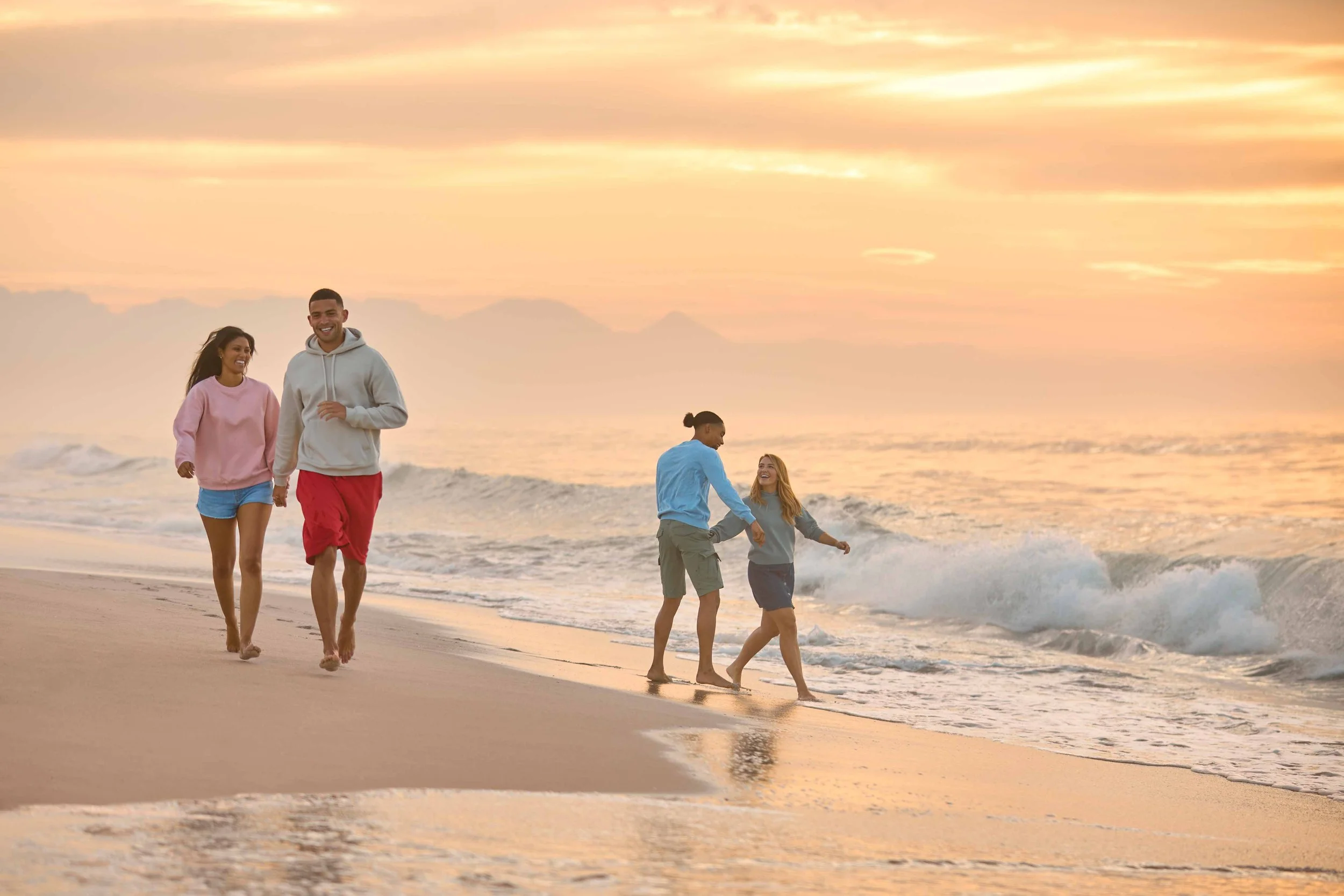 A group of five people, two adults and three children, walking and playing on a sandy beach at sunset, with calm ocean waves and a colorful sky in the background.