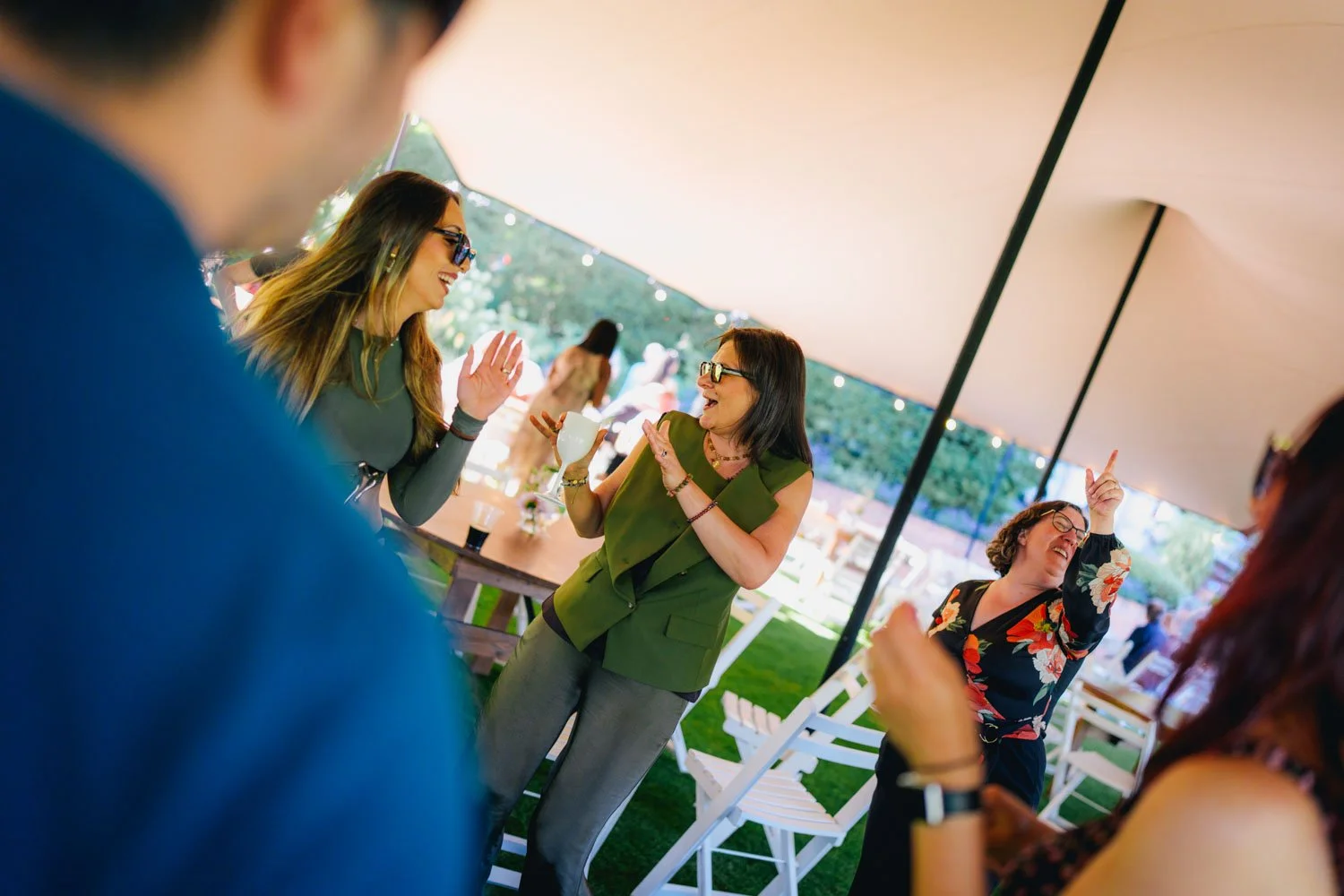 Four women dancing and enjoying themselves at an outdoor party under a large tent, with string lights and white chairs in the background.