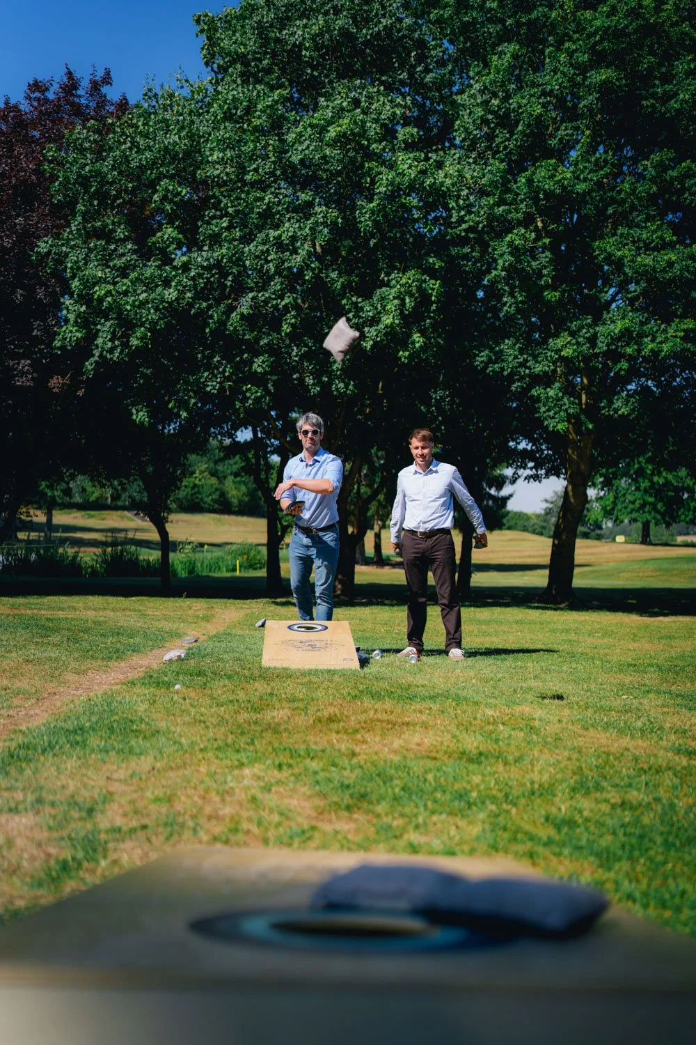 Two men standing on a golf course, one throwing a pillow into the air, with trees in the background.