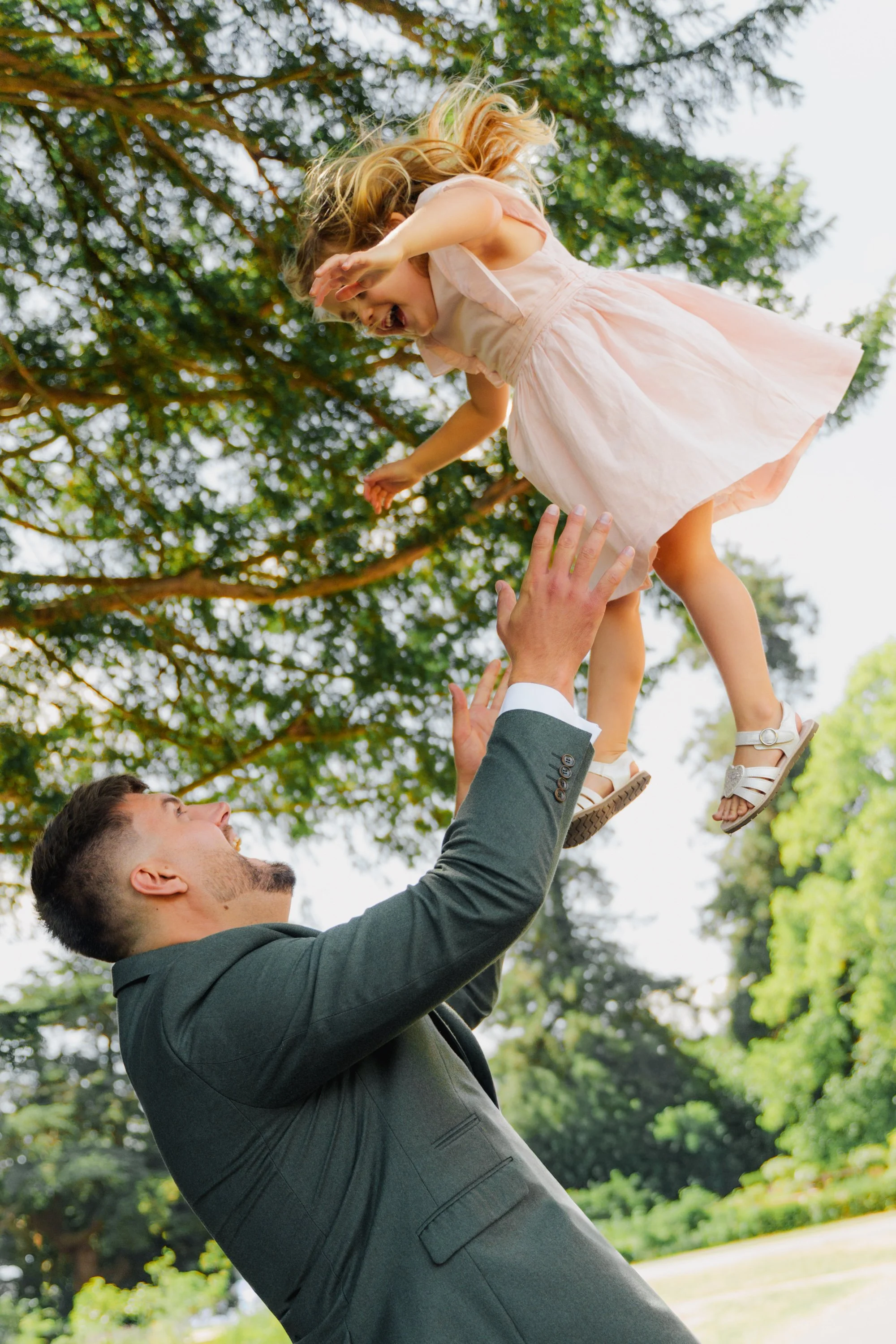 A man in a suit lifting a young girl in a dress high in the air outdoors with trees in the background.