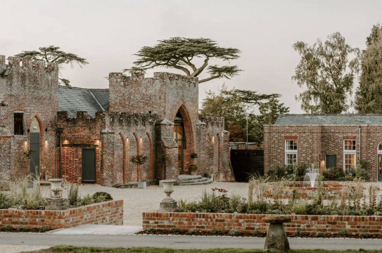 A scenic view of a brick castle-like structure with a tower, arched entrance, and surrounding brick walls. There are trees, garden beds, planters, and a fountain in front of the building.