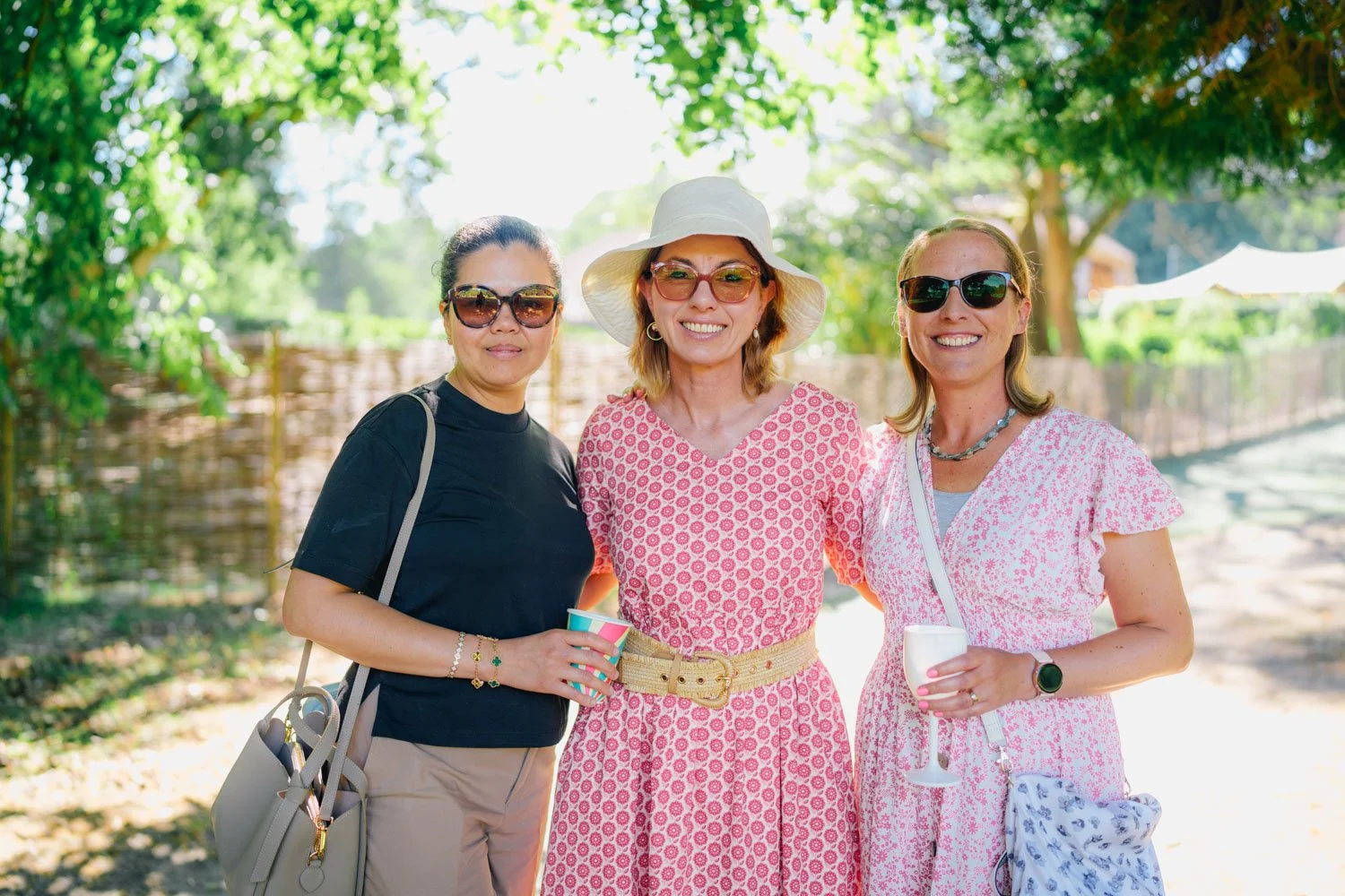 Three women standing outdoors under a canopy of green trees, smiling at the camera, each wearing sunglasses and summery clothing, holding beverages.
