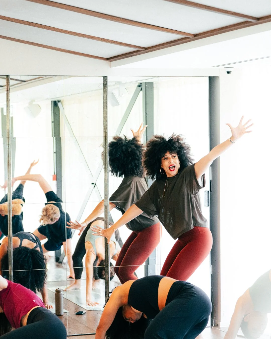 A woman leading a yoga or dance class in front of a mirrored wall, with others in various poses on yoga mats, some bending forward and some in a downward dog position, inside a bright room with large windows.