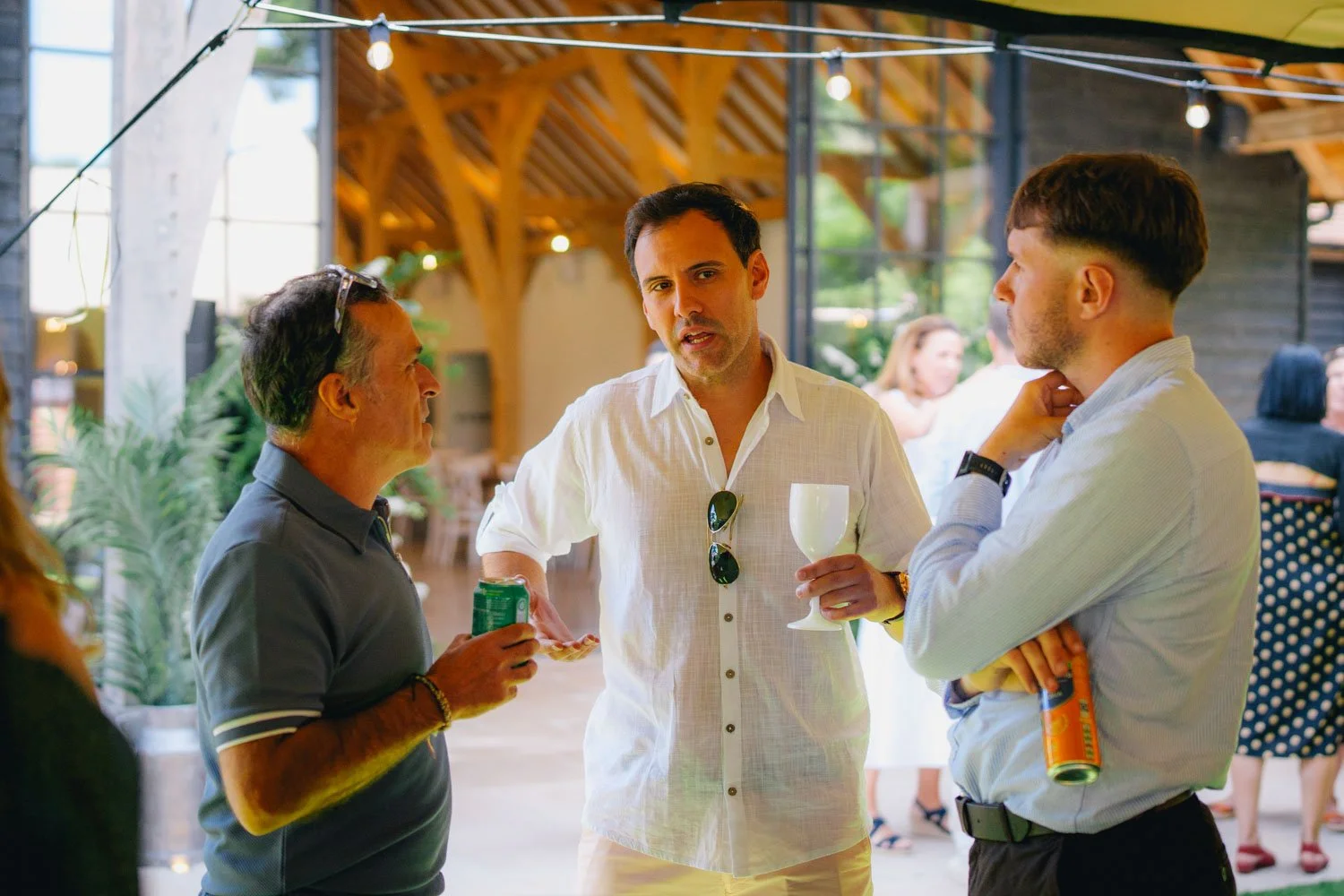 Three men are conversing at a social gathering inside a venue with wooden architecture. The man in the center holds a wine glass, wearing a white shirt with sunglasses hanging from his collar. The man on the left holds a can, wearing a dark striped p