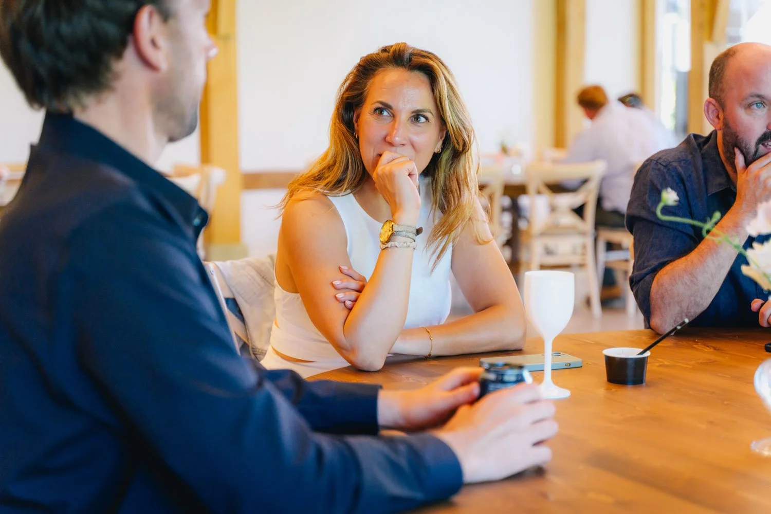 Three people sitting at a wooden table in a restaurant, engaged in conversation; a woman with long blonde hair wearing a white sleeveless top, a man with short brown hair wearing a dark shirt, and another man with a beard wearing a dark shirt, some d
