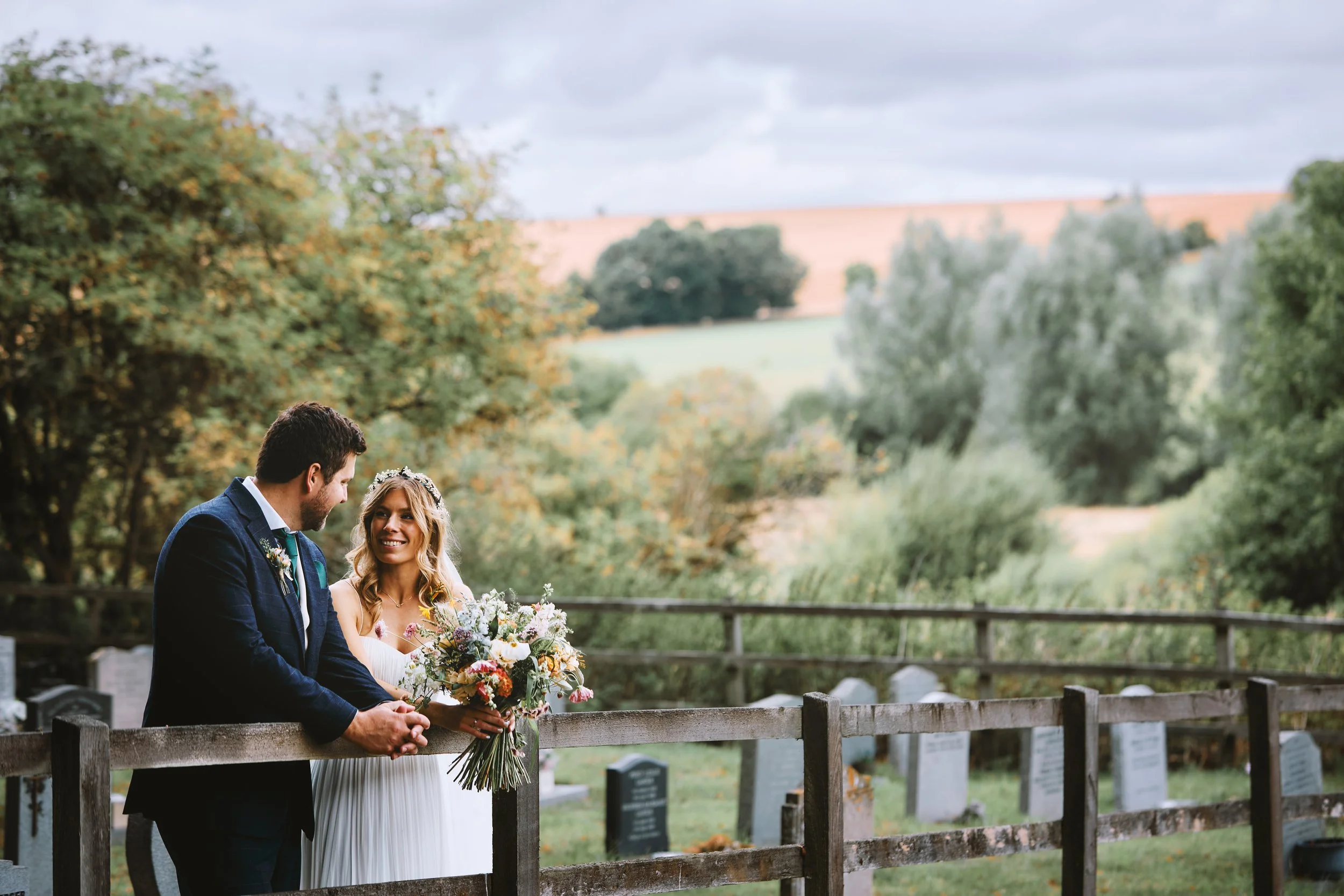 A bride and groom standing together outdoors near a graveyard, holding hands, with trees and a wooden fence in the background during daytime.
