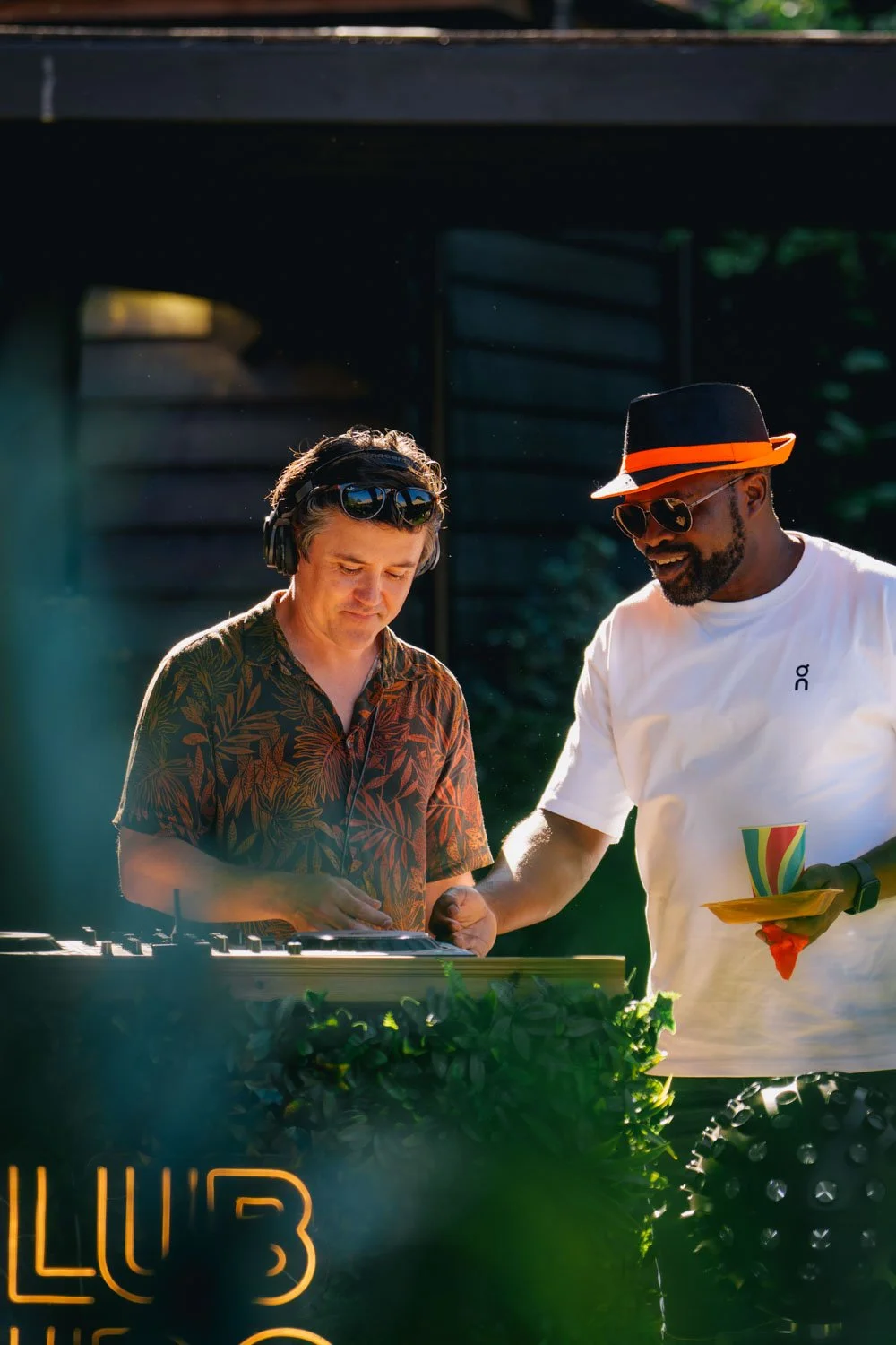 Two men working at a DJ booth outdoors, one wearing headphones and a tropical shirt, the other wearing sunglasses, a hat, and a white shirt, with greenery and a wooden structure in the background.
