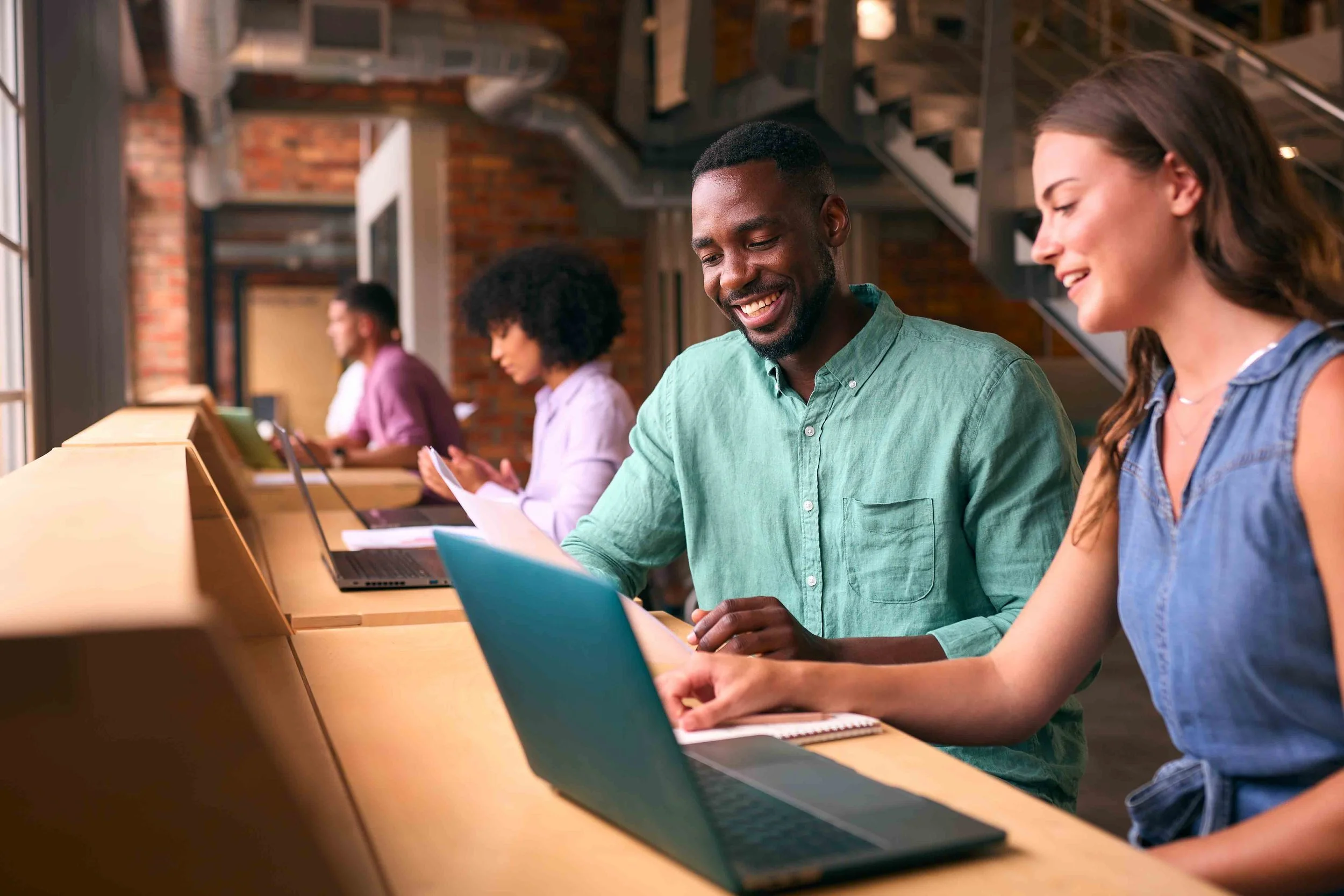 People working on laptops at a wooden counter in a modern, brick-walled workspace with metal staircase.