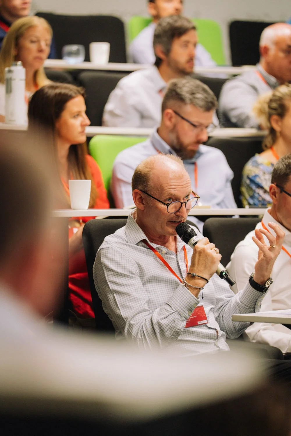 A man with glasses speaking into a microphone at a conference with seated audience members listening, some taking notes, in a lecture hall.