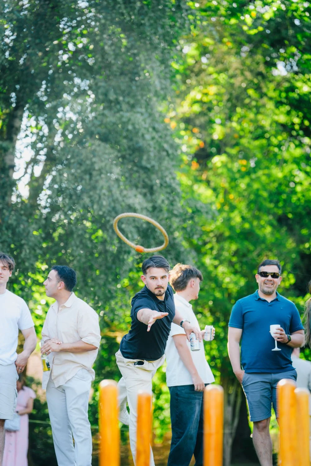 A group of people outdoors at a party, with a man in the center throwing a ring towards a target, while others hold drinks and chat, surrounded by green trees and sunlight.