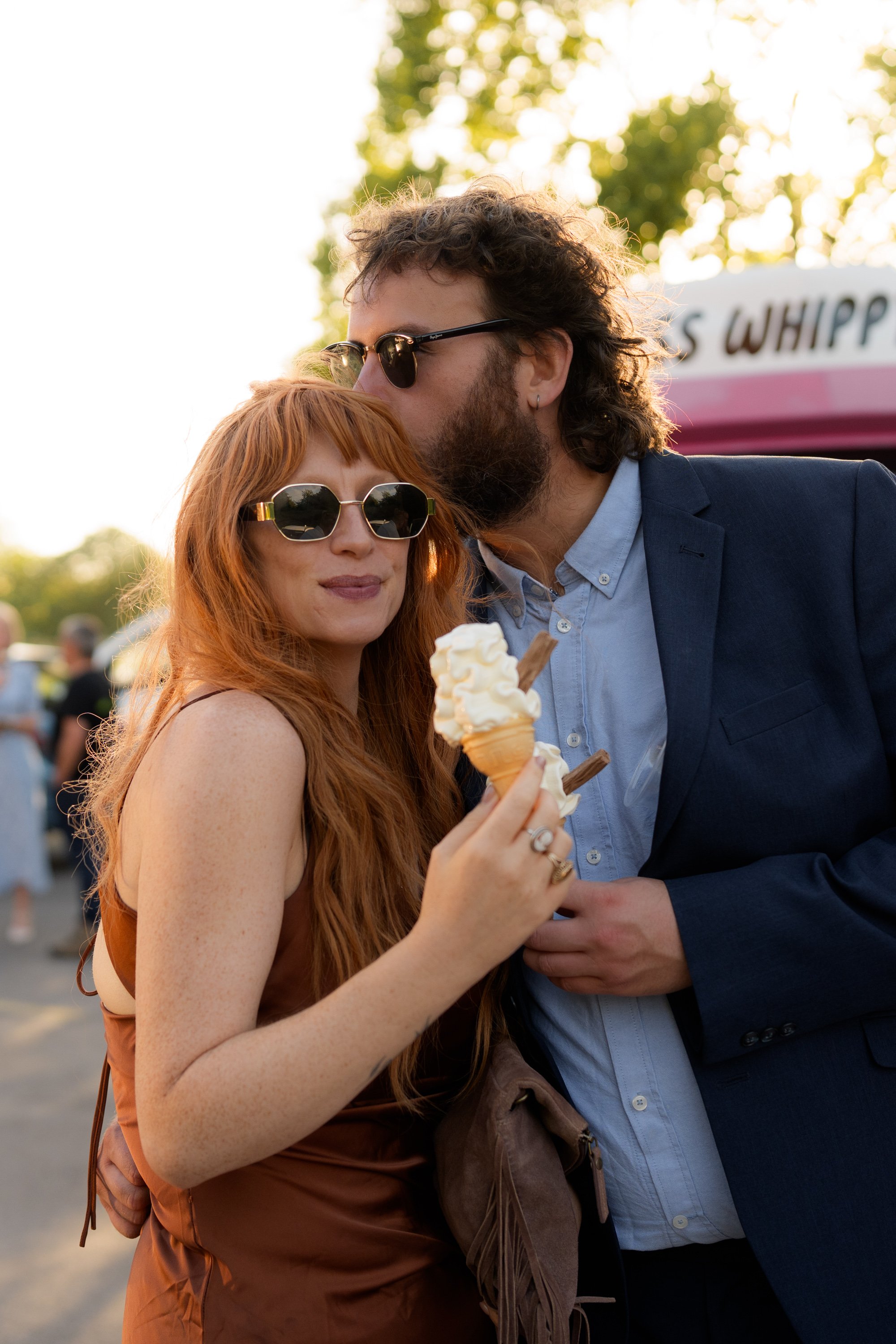 A man with long curly hair, wearing sunglasses and a blue suit, kisses a woman with long red hair and sunglasses, holding an ice cream cone, outdoors during daylight with trees in the background.