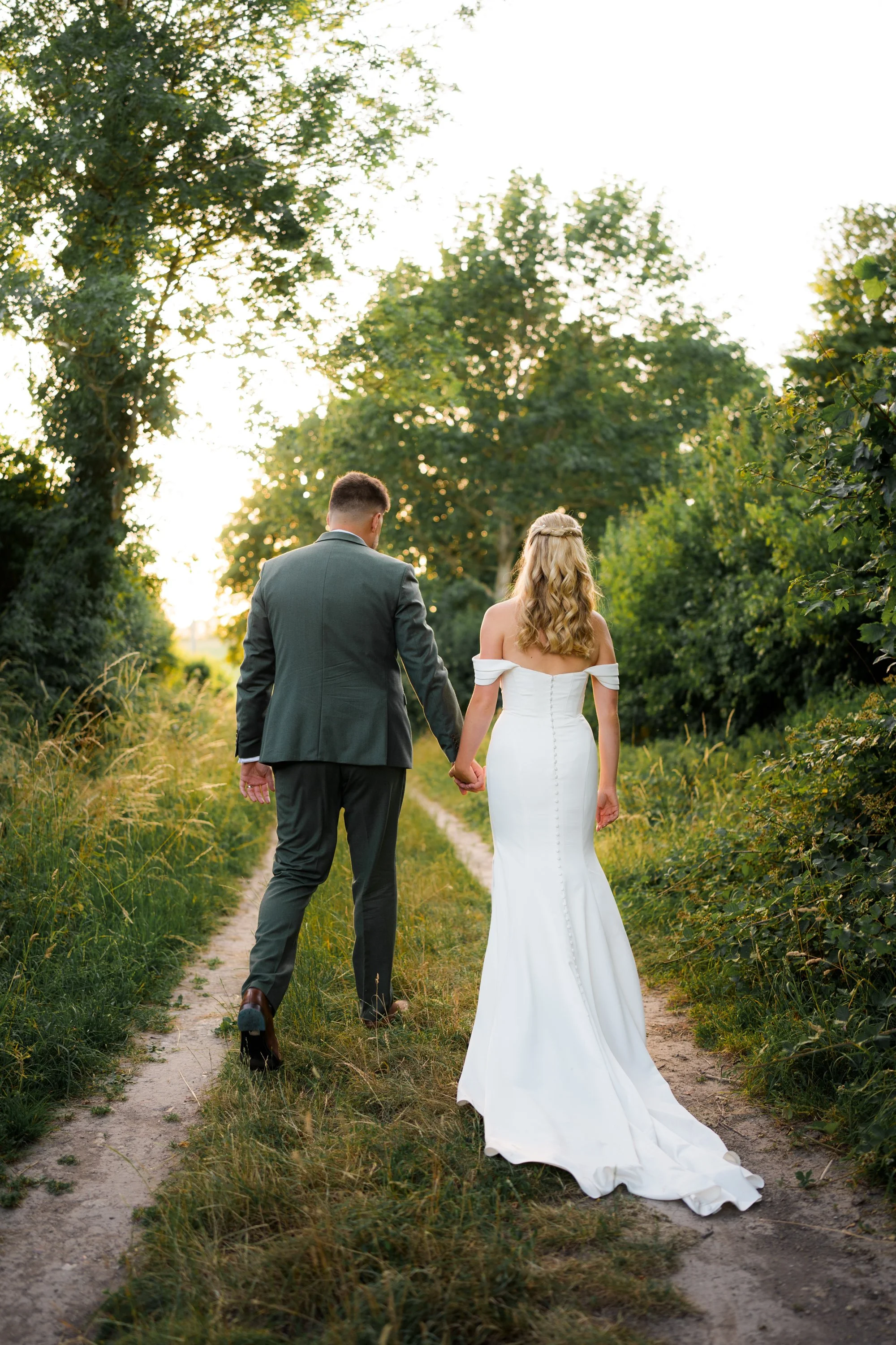 A bride and groom holding hands, walking on a dirt path surrounded by lush trees and greenery during sunset.
