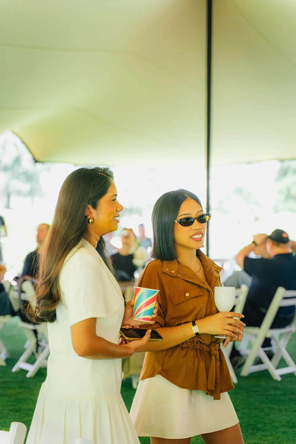 Two women at an outdoor gathering, one in a white dress holding a striped cup and phone, the other in a brown top with sunglasses holding a white candle holder, with seated guests in the background.
