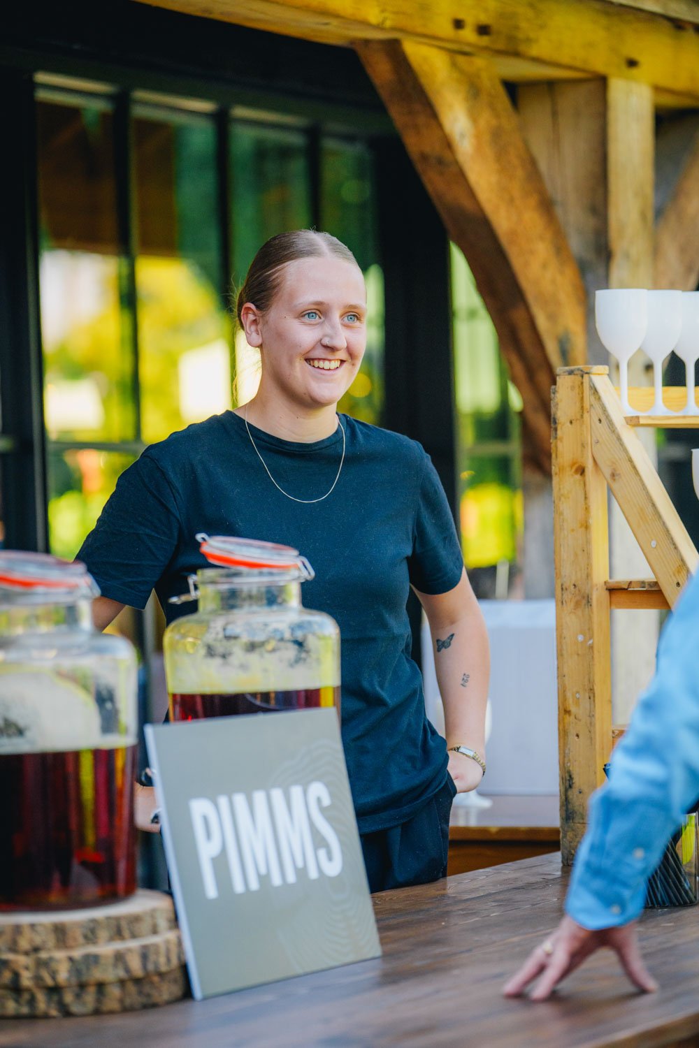 A woman with blonde hair smiling behind a bar counter with jars labeled "PIMMS"; she is wearing a black T-shirt and standing outdoors with a wooden structure and greenery in the background.