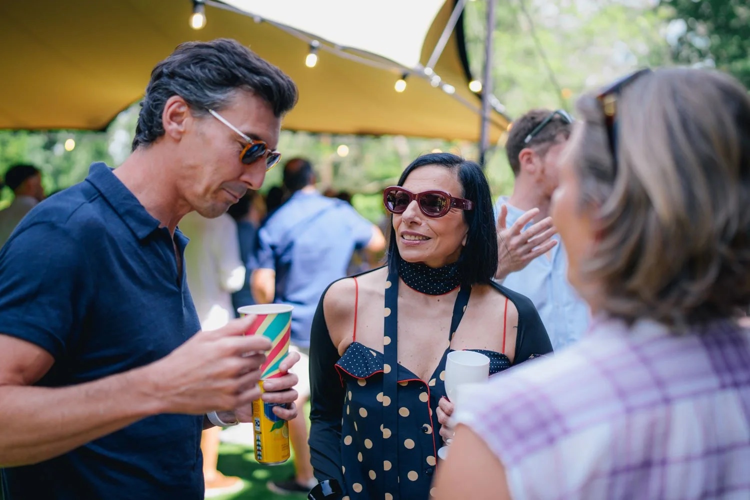 Three people at an outdoor party are talking and holding drinks under a yellow canopy with string lights.