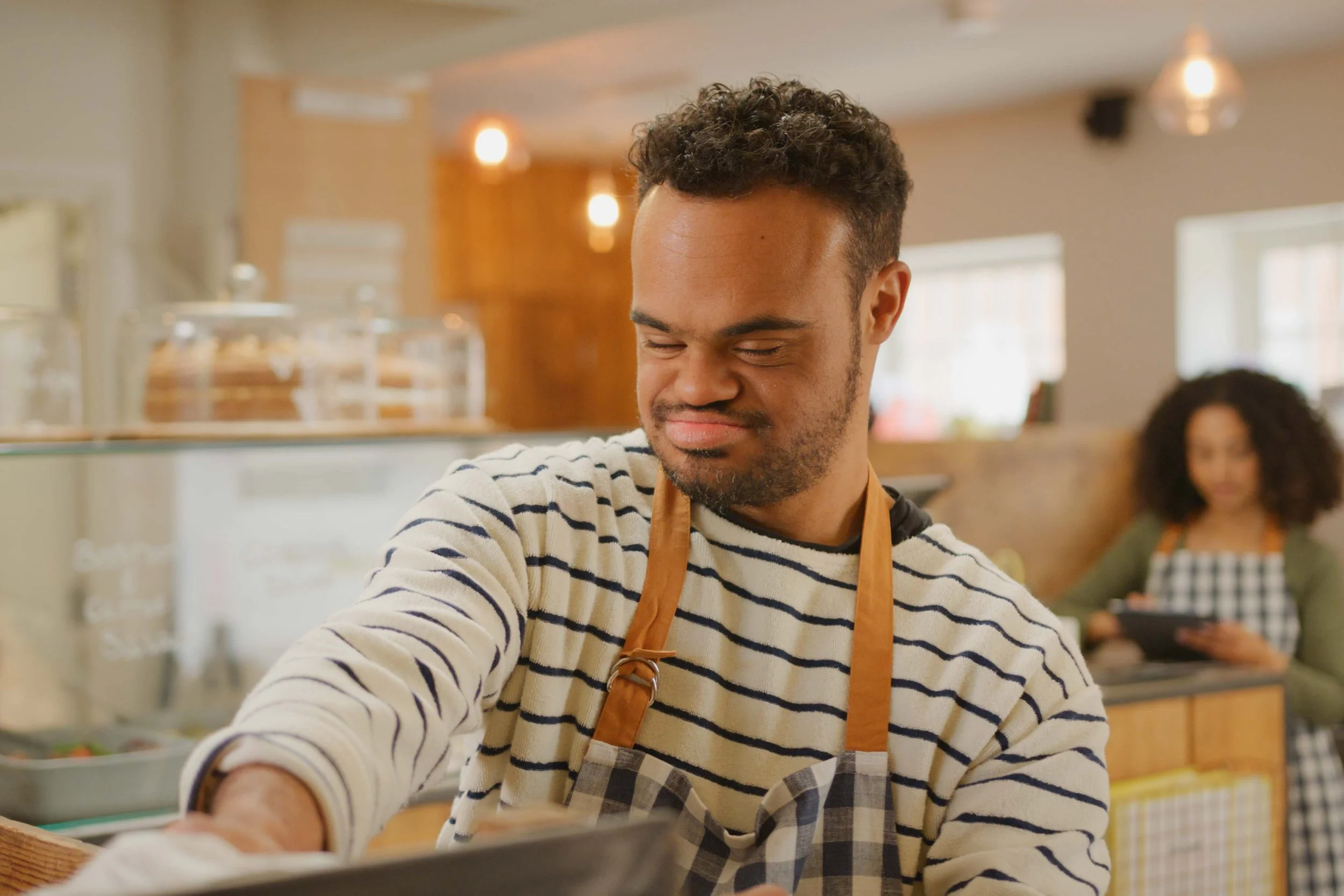 A young man with curly hair and a beard, wearing a striped shirt and a brown apron, smiling while working in a cafe or restaurant, with a blurred background of another woman working nearby.