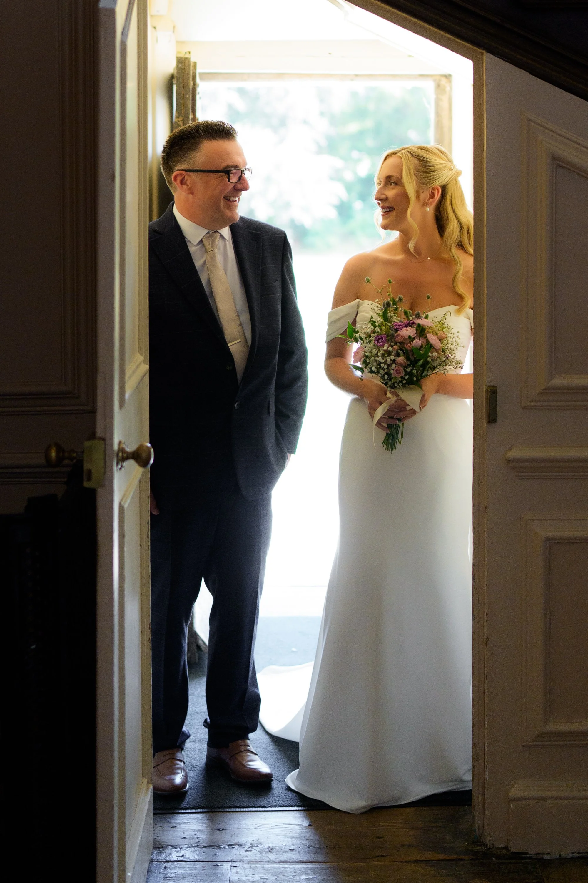 A bride holding a bouquet of flowers smiling at a man in a suit as they stand in an open doorway.