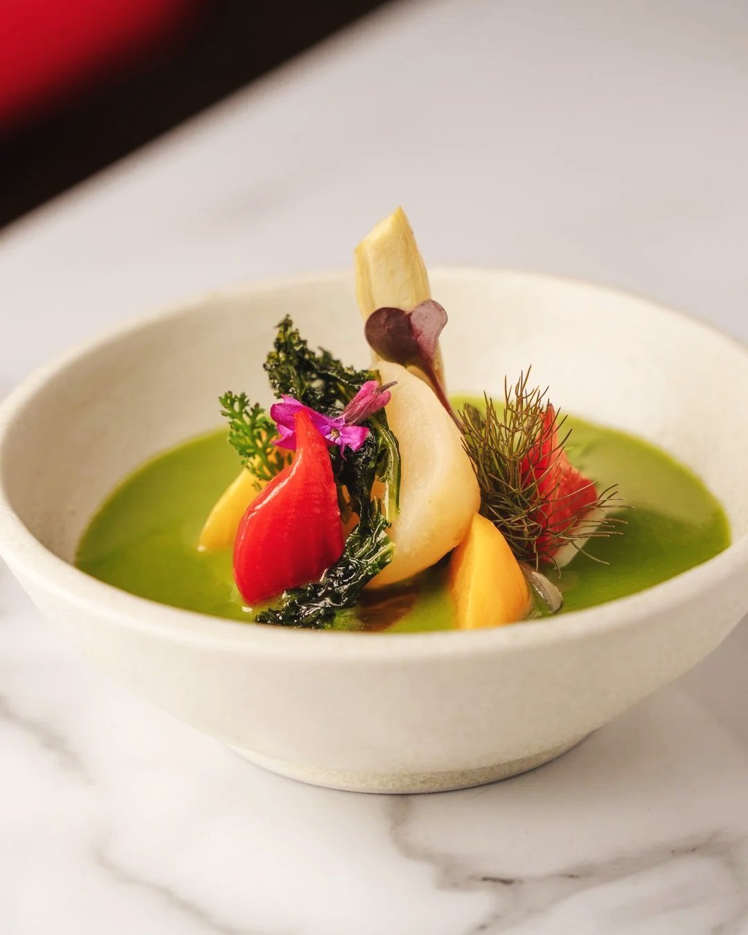 A bowl of green soup garnished with small red, yellow, and purple vegetables, edible flowers, herbs, and a piece of root vegetable.