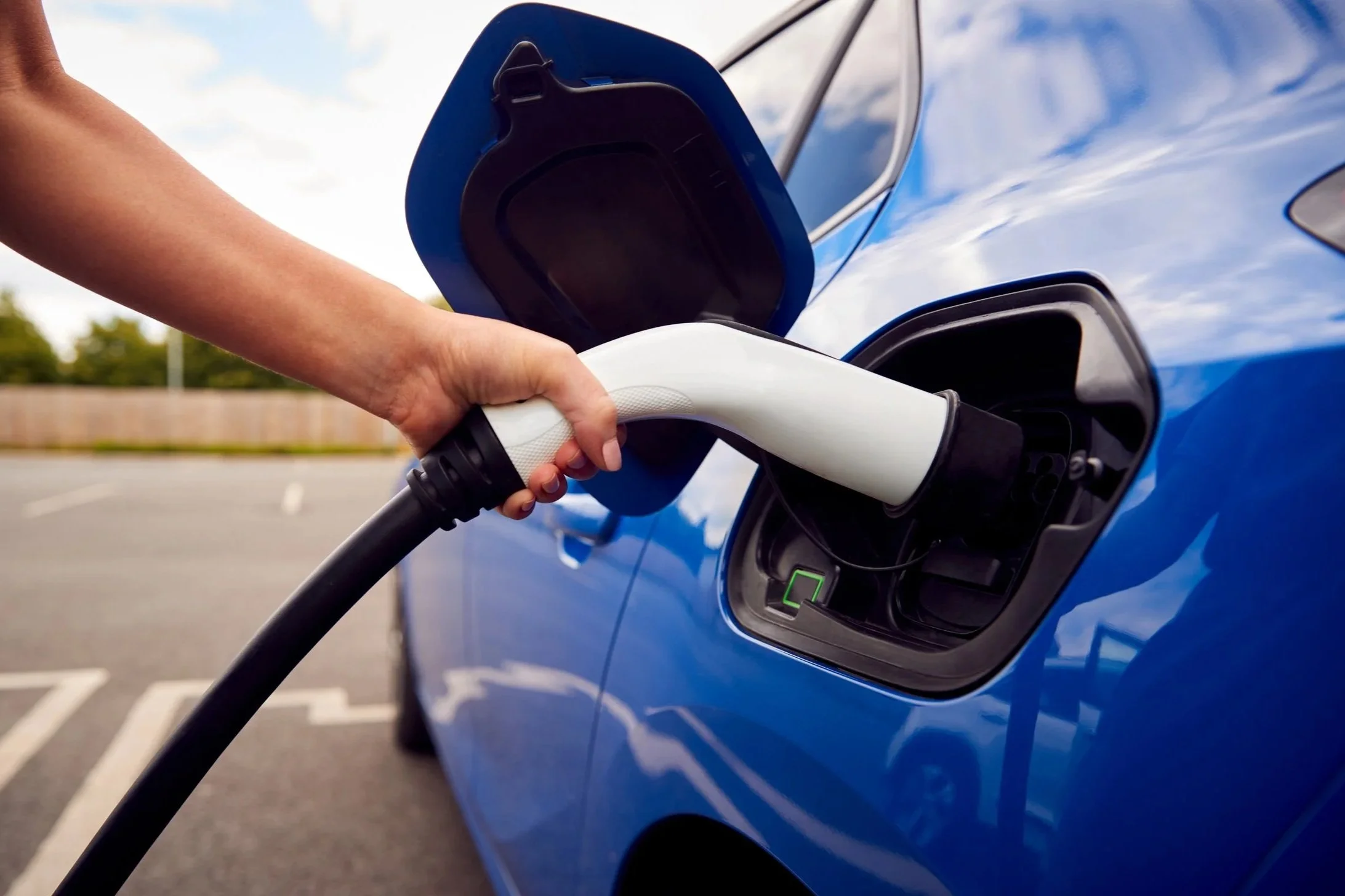 A person is plugging in a charging cable into an electric vehicle's charging port on a blue car, with a parking lot and cloudy sky in the background.
