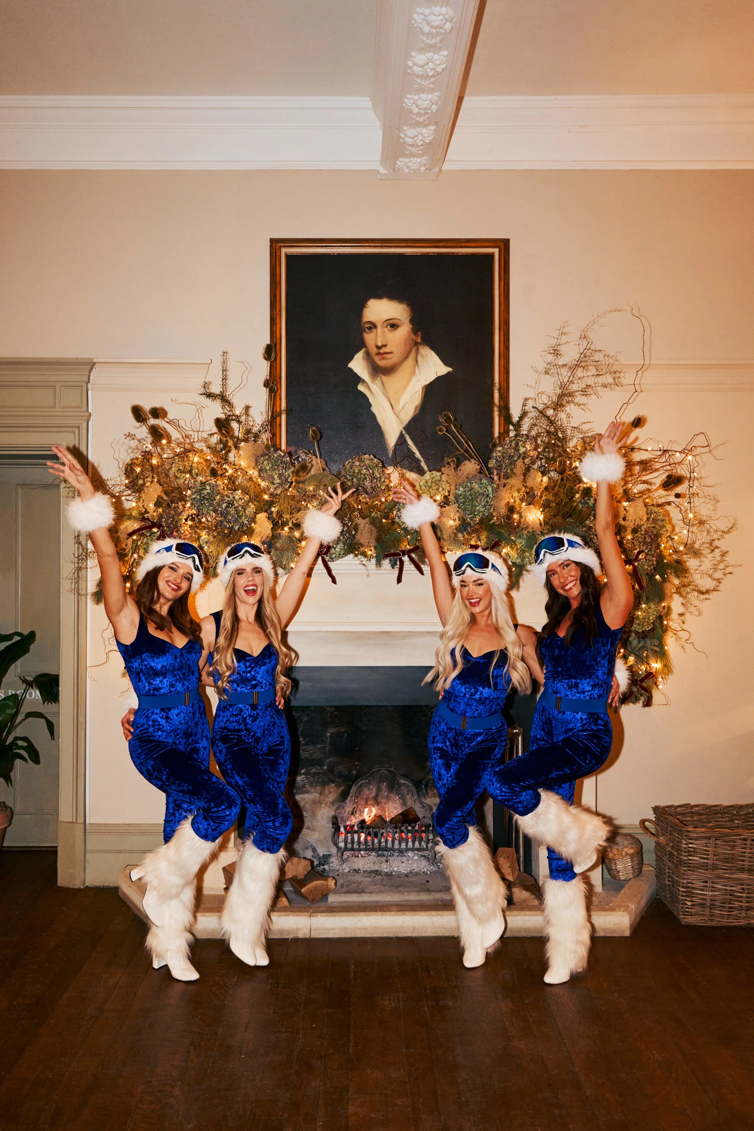 Four women dressed in matching blue velvet holiday costumes with white faux fur boots and accessories, posing cheerfully in front of a fireplace decorated with a large, illuminated garland and a classical portrait painting above the mantel.