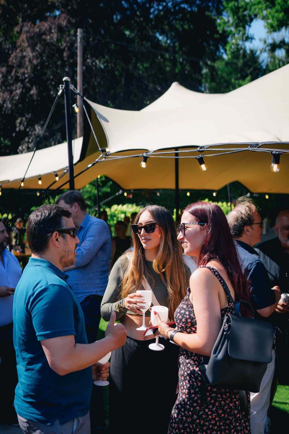 A group of people gathered outdoors at a social event, talking and holding drinks under large white umbrellas, with trees and string lights in the background.