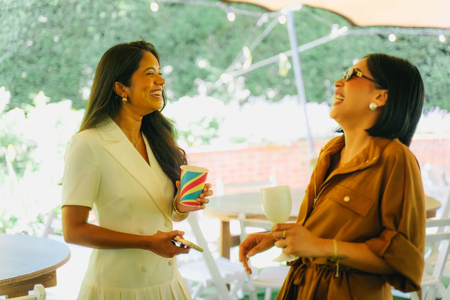 Two women are smiling and enjoying a conversation at an outdoor gathering, holding cups and standing under a canopy with string lights, with a green, leafy background.