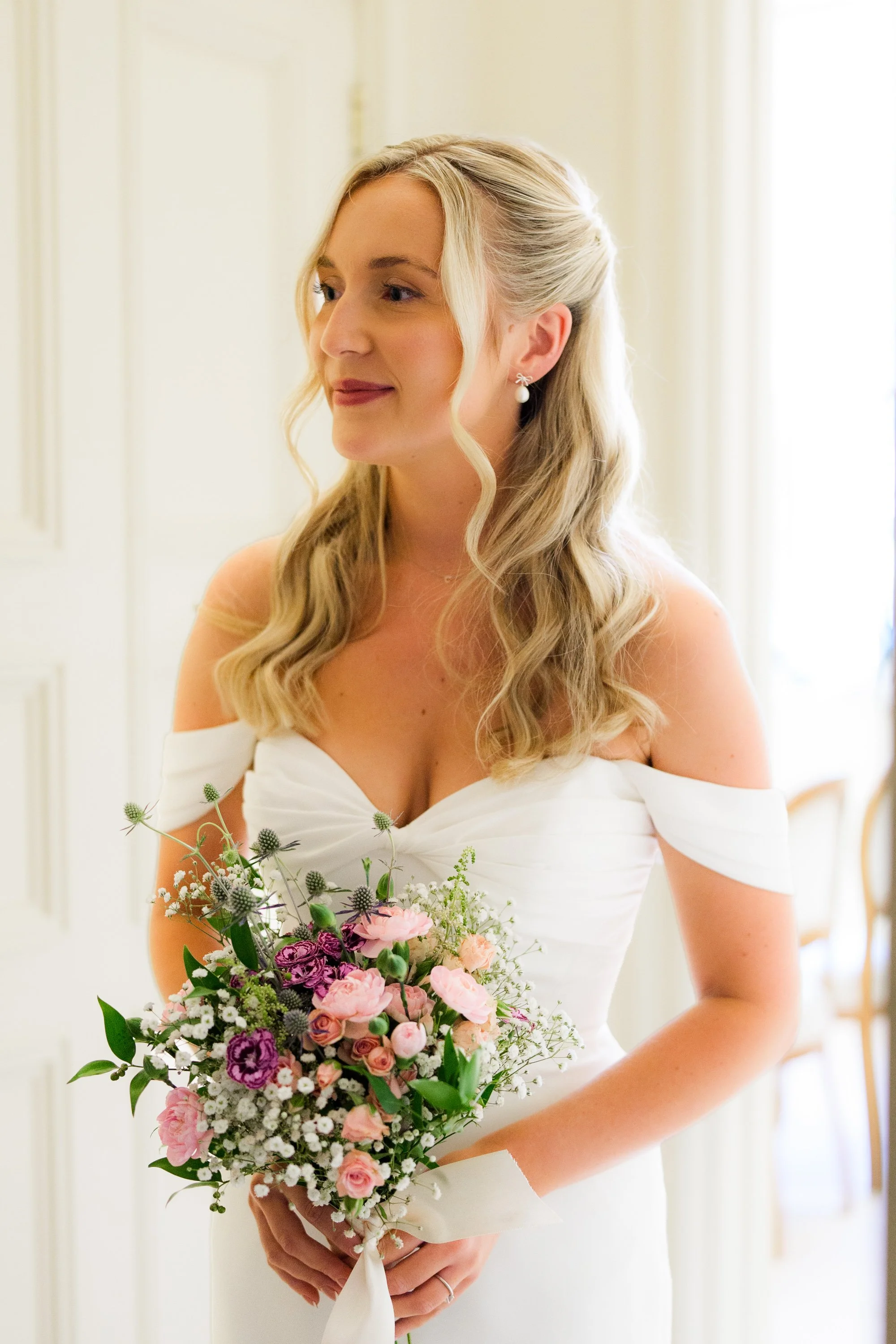 A bride with blonde hair styled in loose waves, wearing pearl earrings and a white off-the-shoulder wedding dress, holding a colorful bouquet of pink, purple, white, and green flowers.