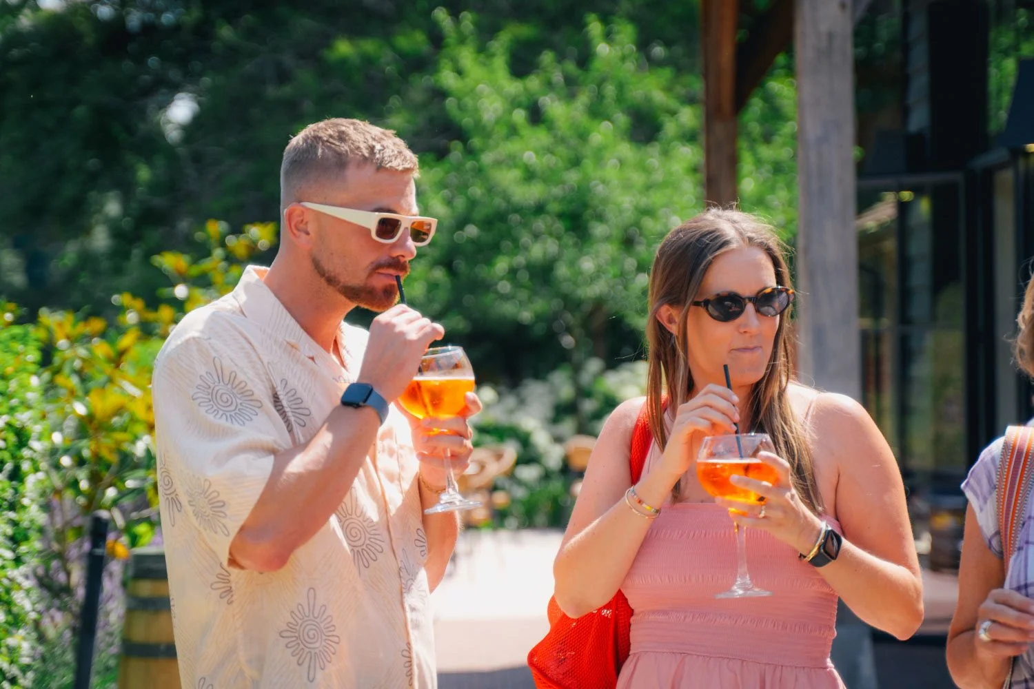 Two people drinking orange-colored cocktails with black straws outdoors on a sunny day. The man is wearing a light-colored patterned shirt and white sunglasses, and the woman is wearing a pink dress with sunglasses. Green trees and a wooden structure
