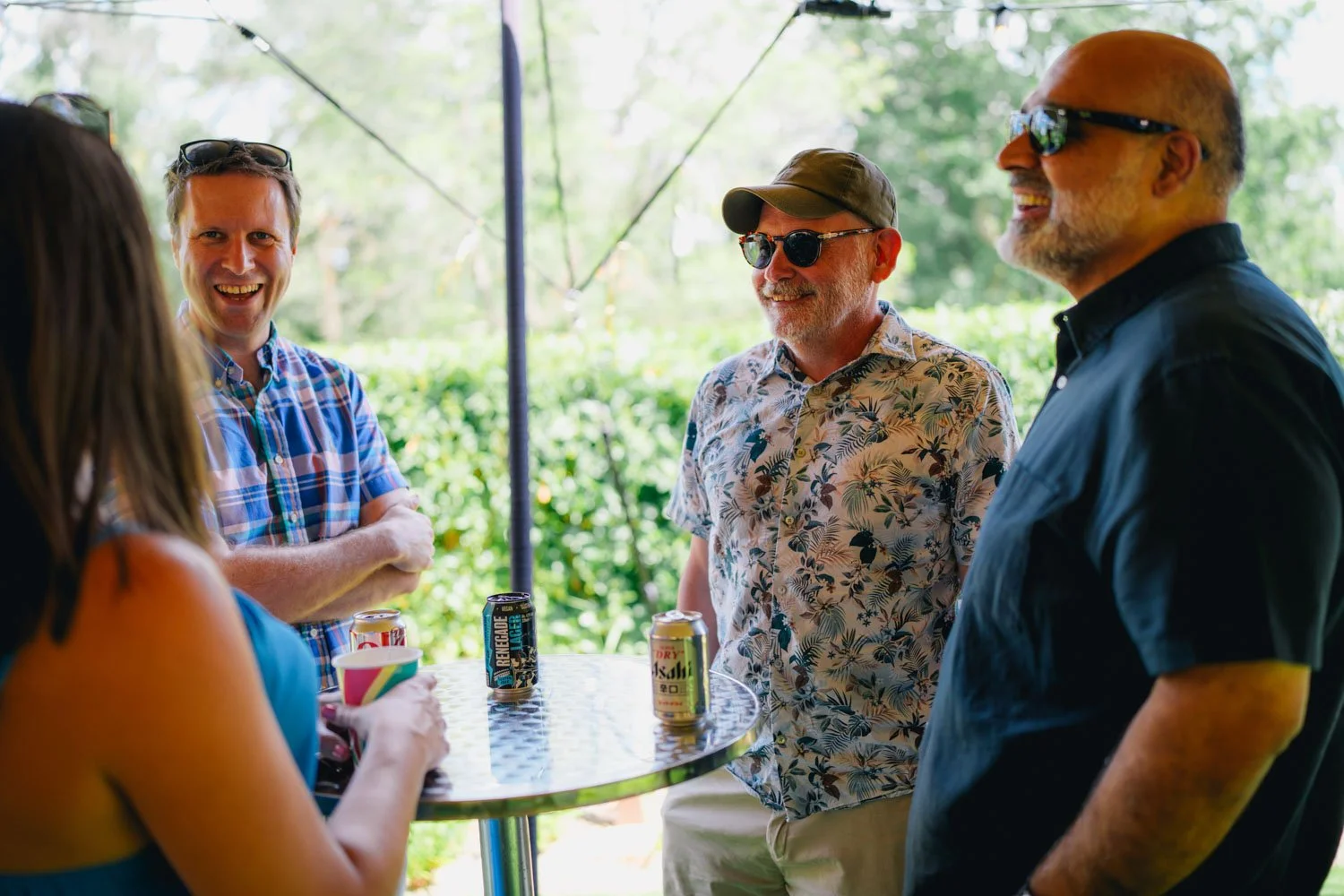 Four people are standing around a metal outdoor table with drinks, talking and smiling. The background shows greenery and trees.