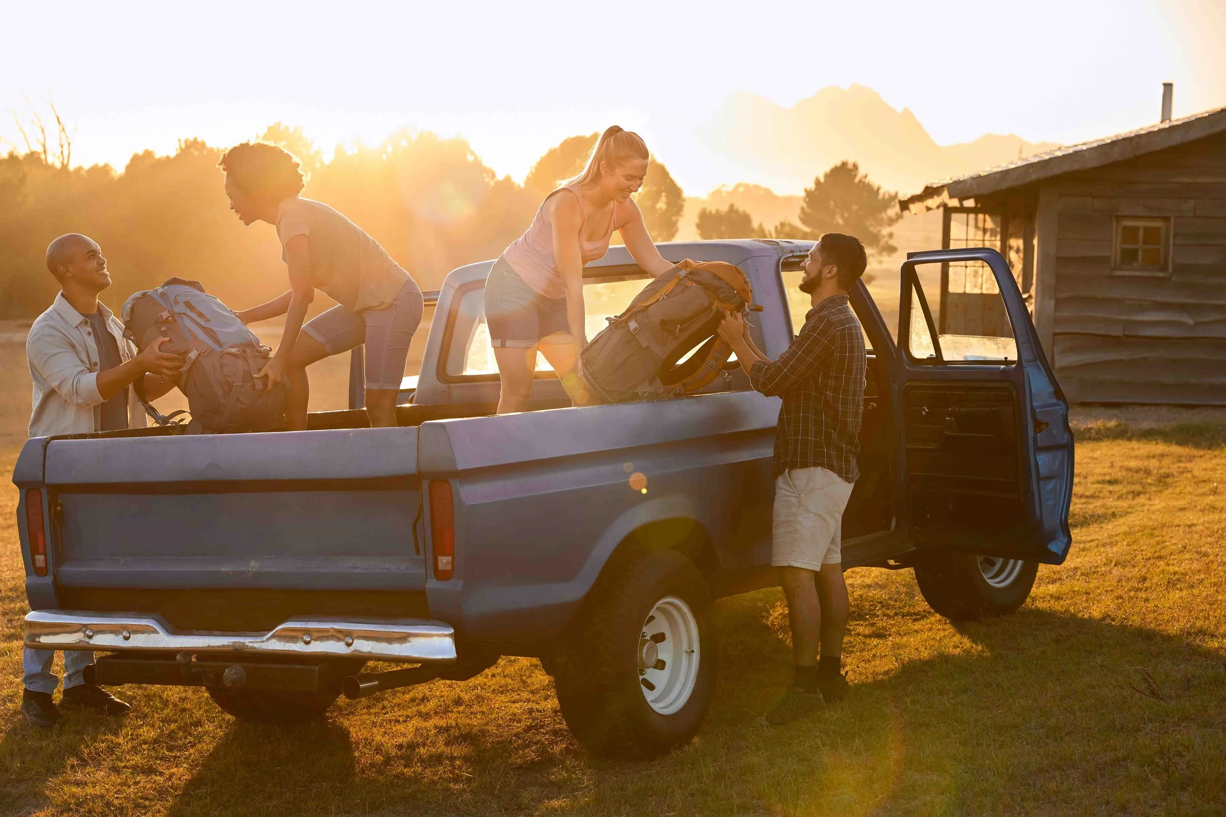 Four young adults loading hiking backpacks into the back of a pickup truck during sunset in a rural area with a wooden shed in the background.